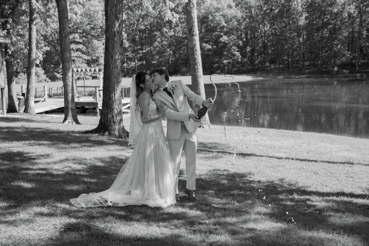 Black and white photo of a bride and groom sharing a kiss outdoors near a serene lake surrounded by tall trees.