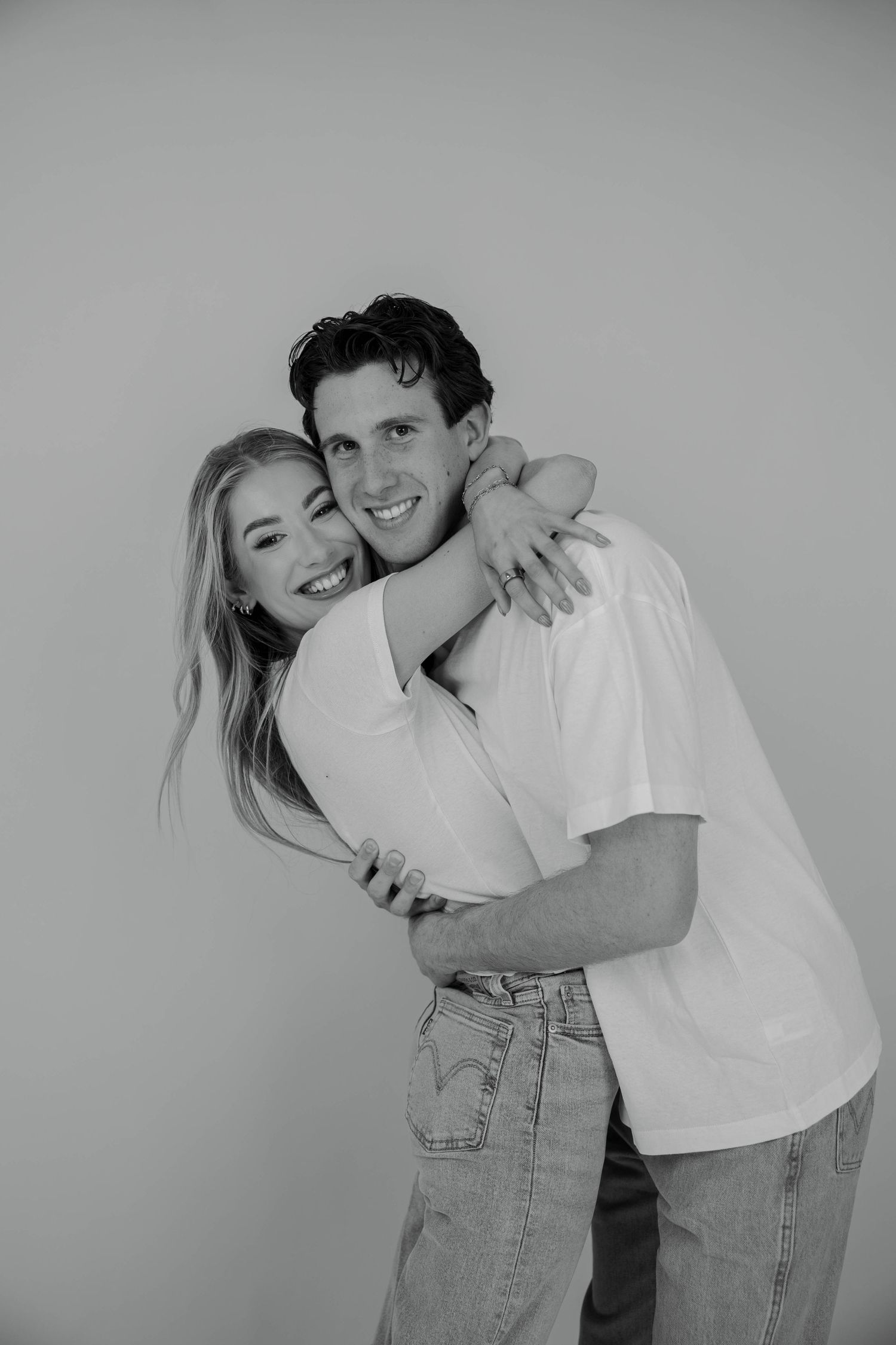 Black and white photo of a smiling couple hugging, woman with blonde hair embracing man in white t-shirt and jeans.