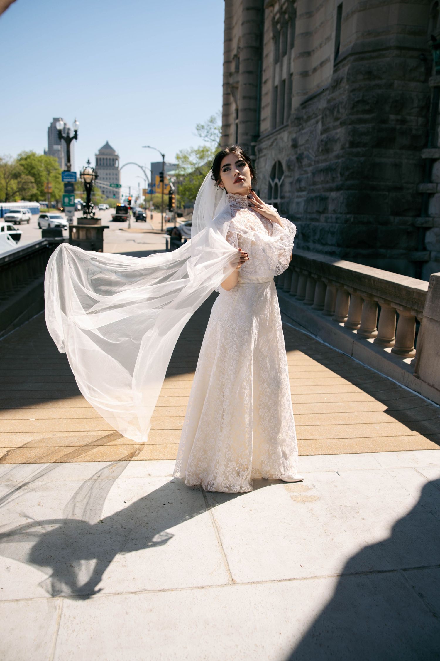 Bride in long-sleeve lace wedding dress with veil flowing in wind stands on sunny stone balcony.
