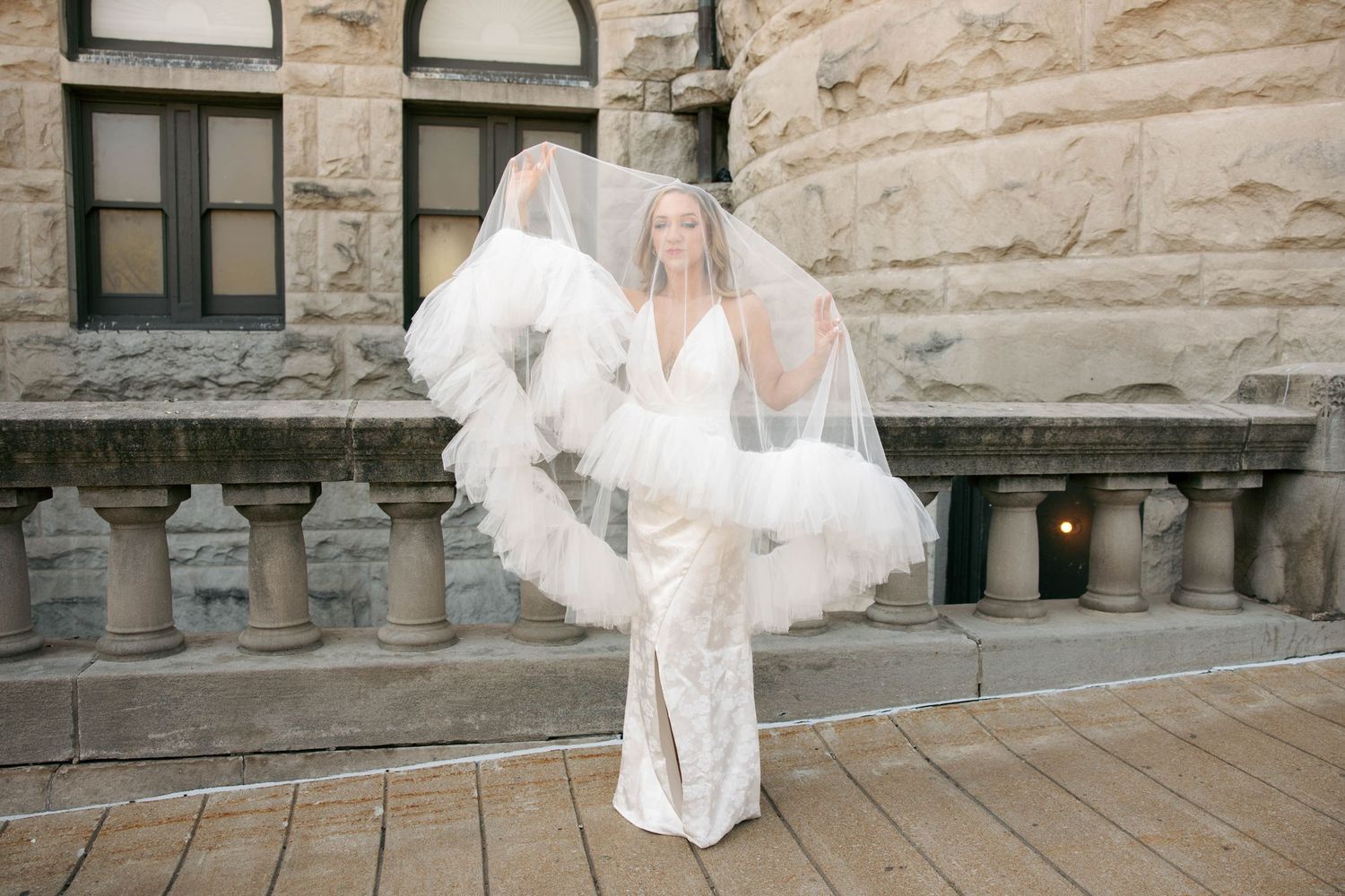 Bride in fitted lace wedding gown with flowing feather veil outdoors near stone building.