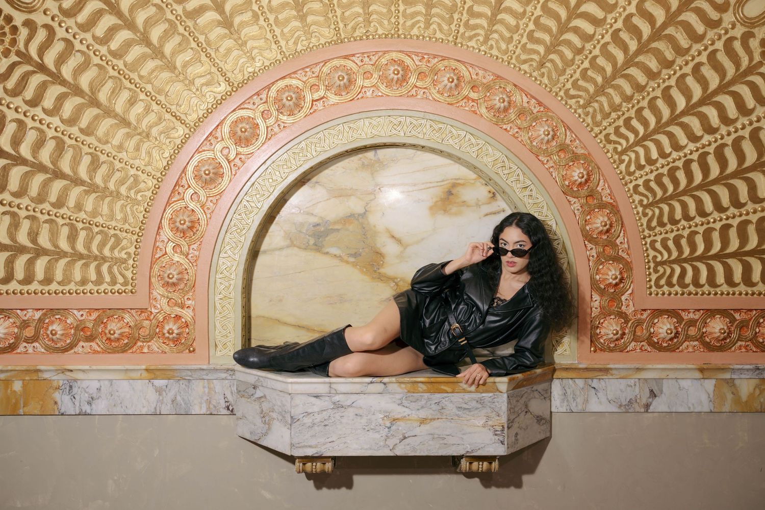 Woman in black leather jacket poses in ornate golden arch niche with marble ledge and decorative molding.