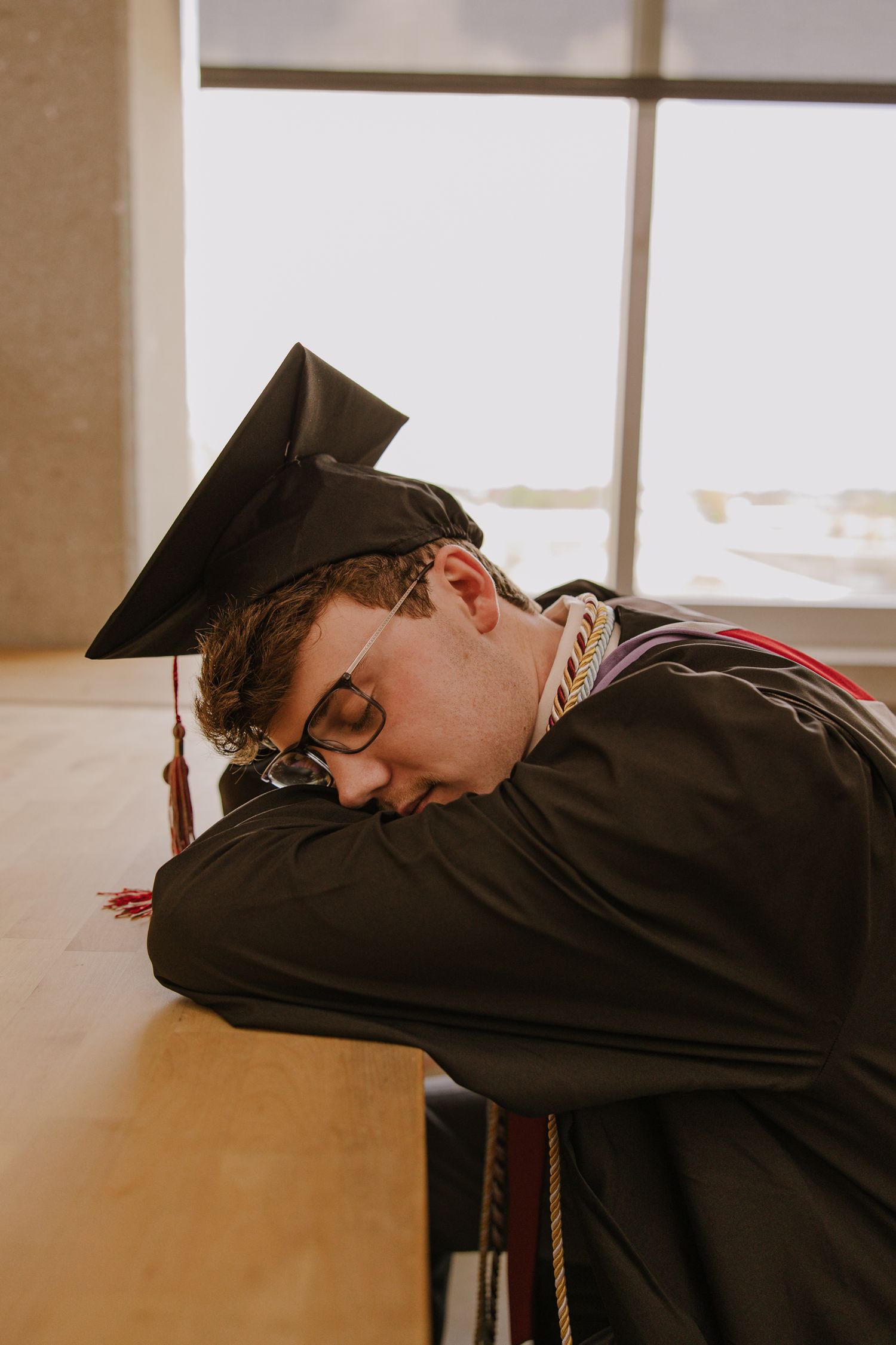 Graduate in cap and gown rests head on desk in artistic graduation photo.