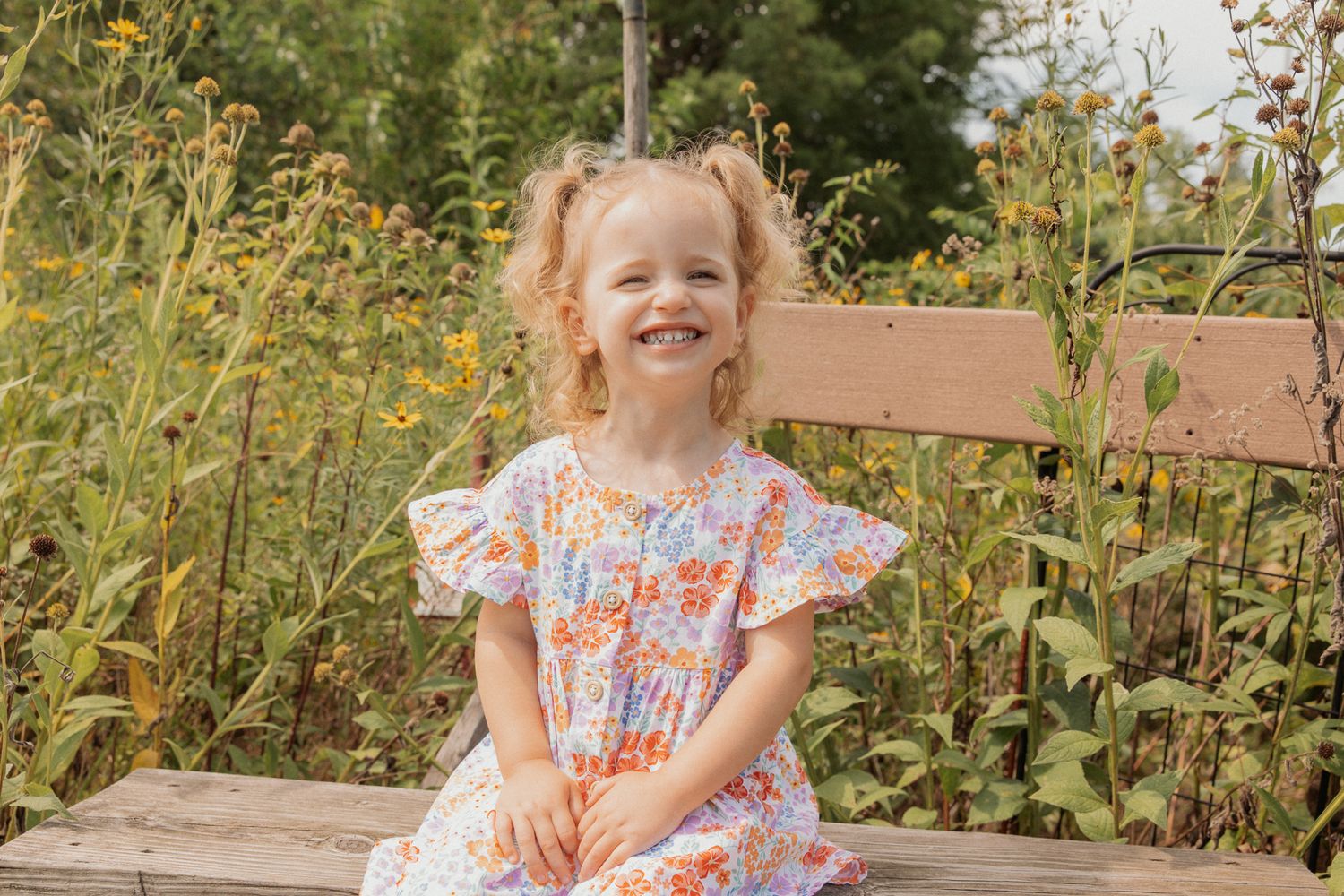 Young girl in floral dress poses playfully on wooden bench surrounded by yellow wildflowers.