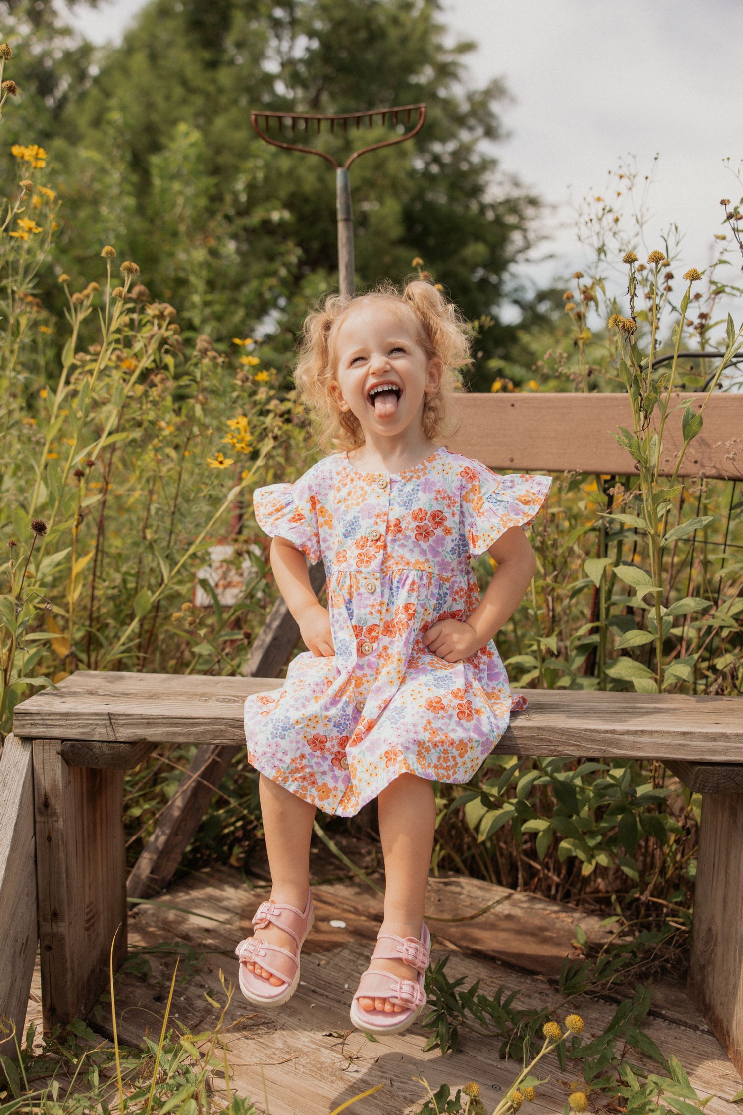 Child in floral dress sits joyfully on wooden bench among blooming flowers.