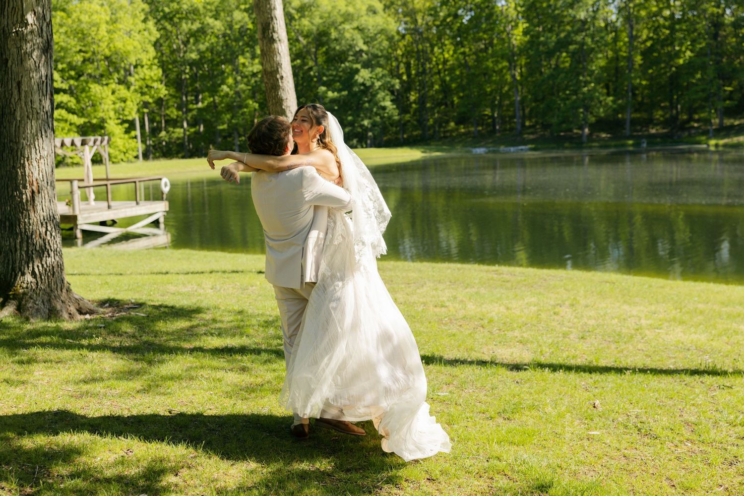 Bride and groom embracing by a tree near a serene pond in a lush green outdoor wedding setting.