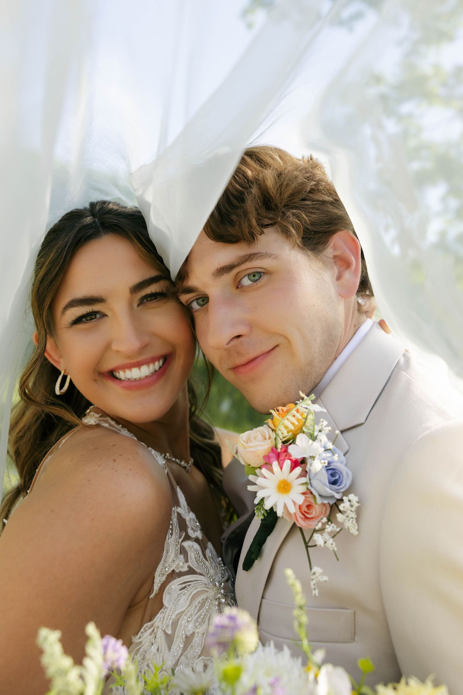 A smiling bride and groom pose closely together under a white veil, with colorful wedding flowers in the foreground.
