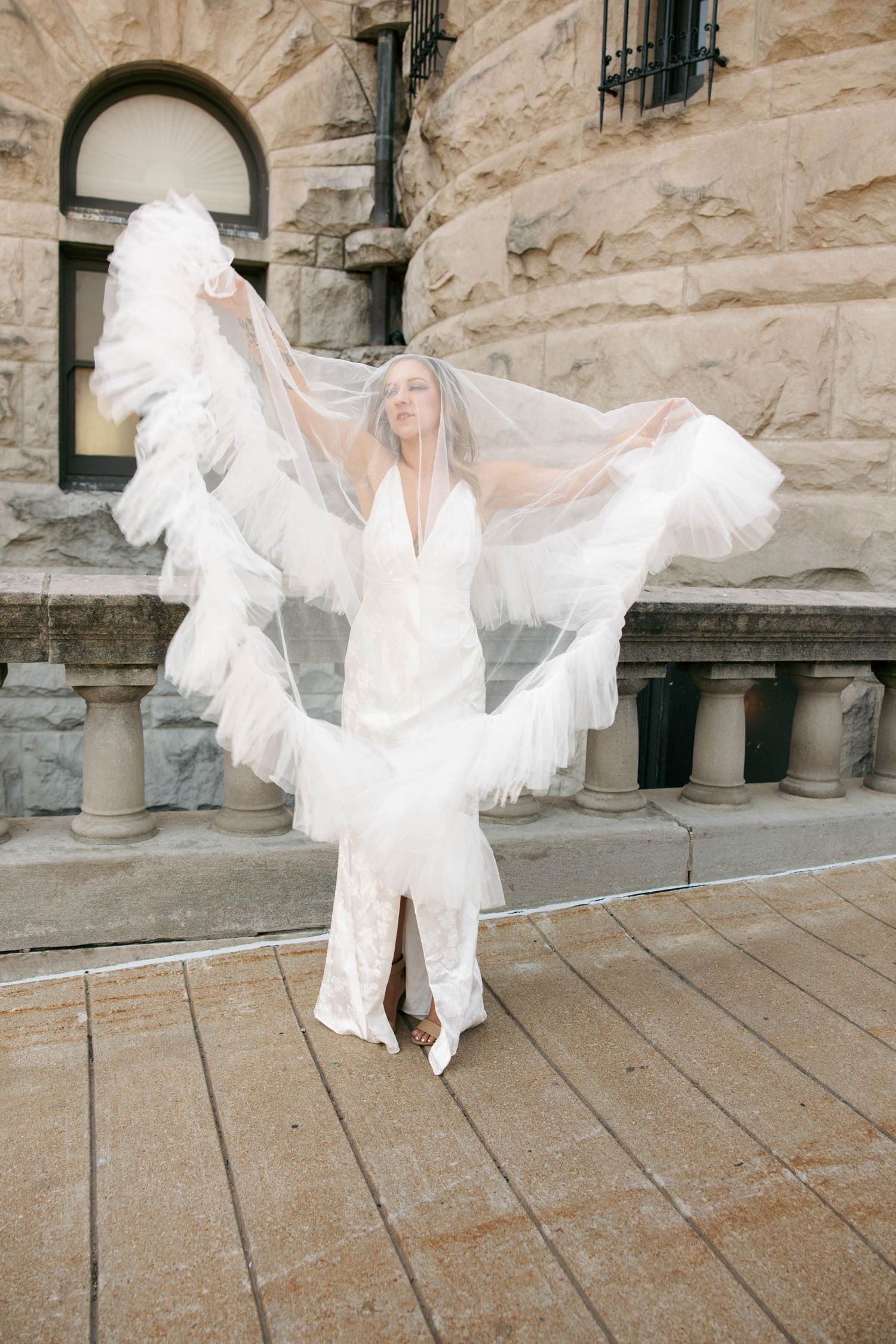 Bride in sleek white gown lifting voluminous ruffled veil with both hands in front of historic stone building.