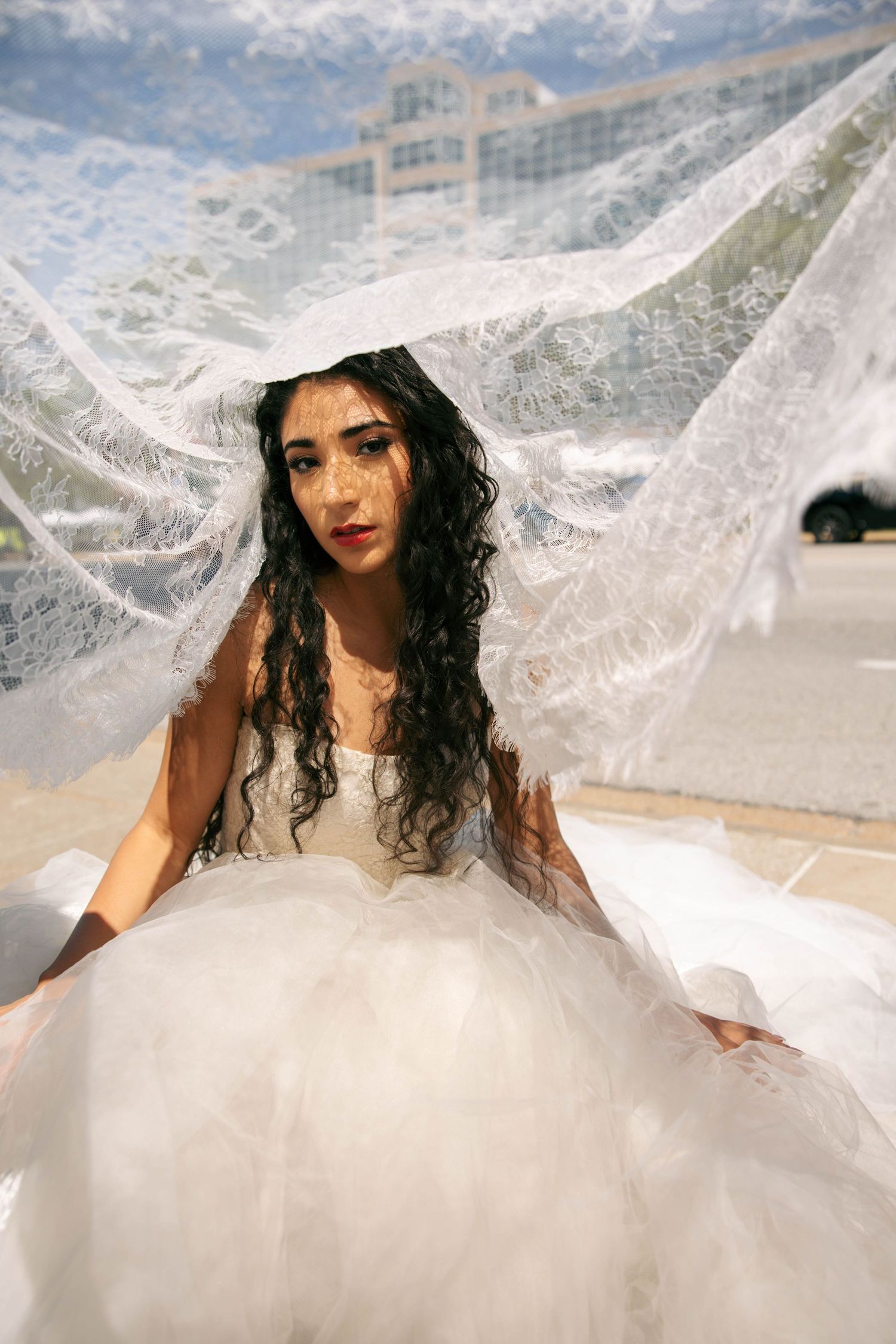 Bride in white ball gown and lace veil billowing overhead poses outdoors near modern building.