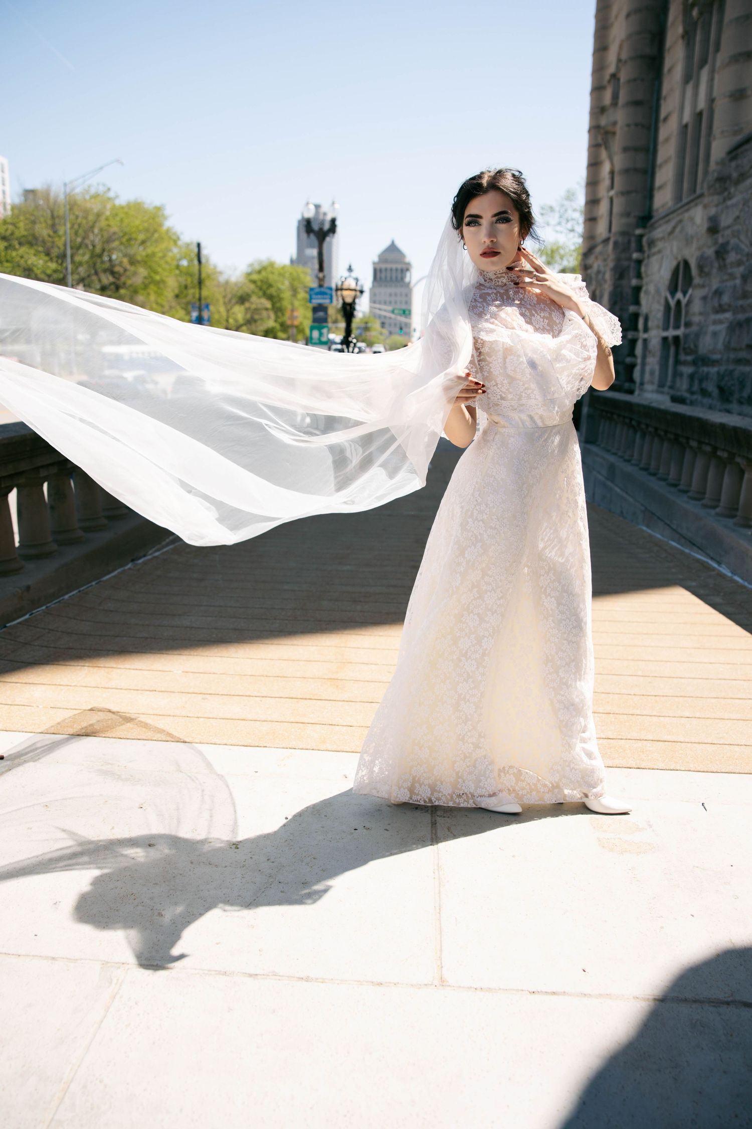 Elegant bride in lace gown with billowing veil stands on sunny outdoor steps near historic building.