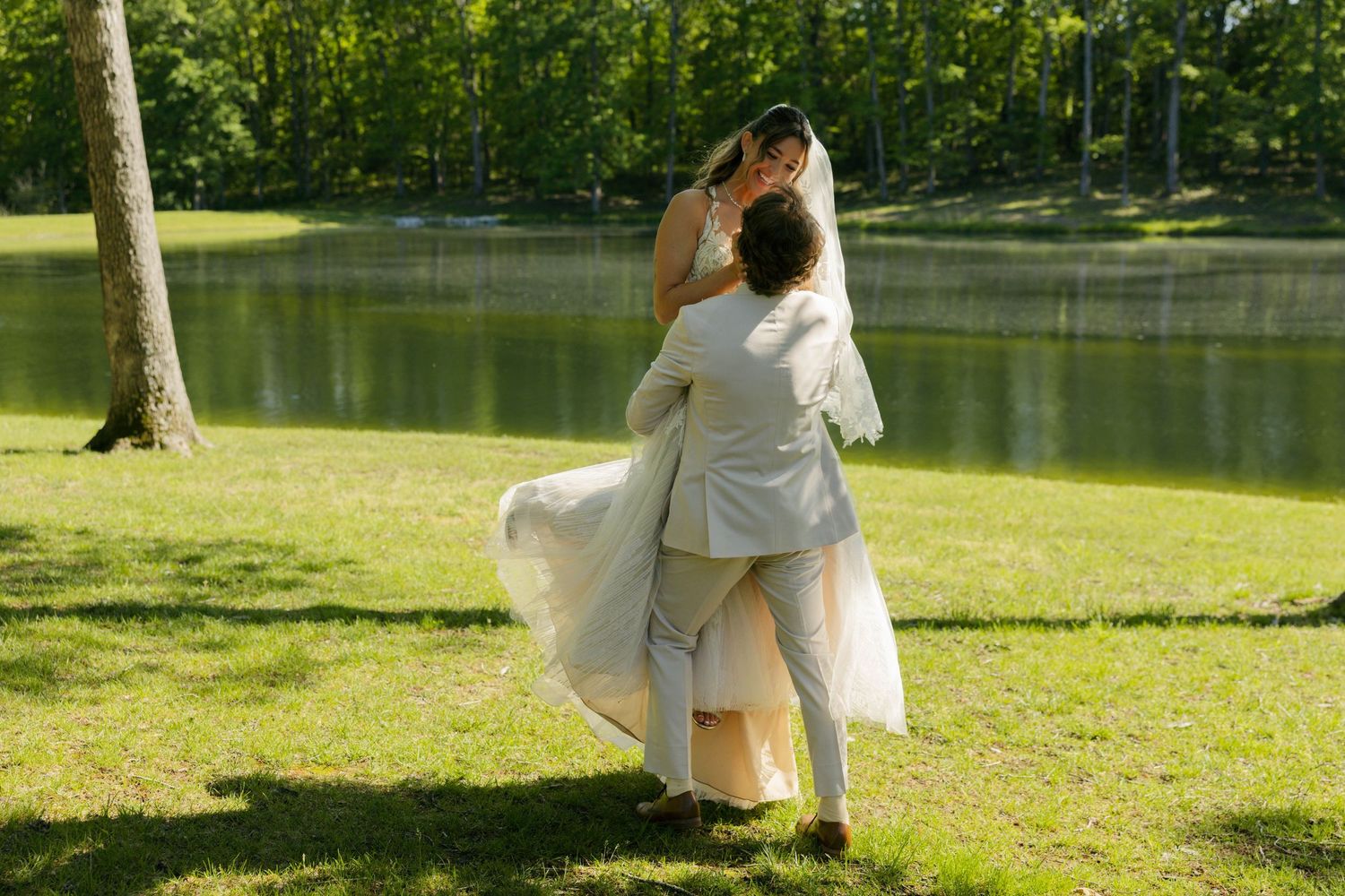 Bride and groom dancing on green lawn by a serene pond surrounded by lush trees on their wedding day.