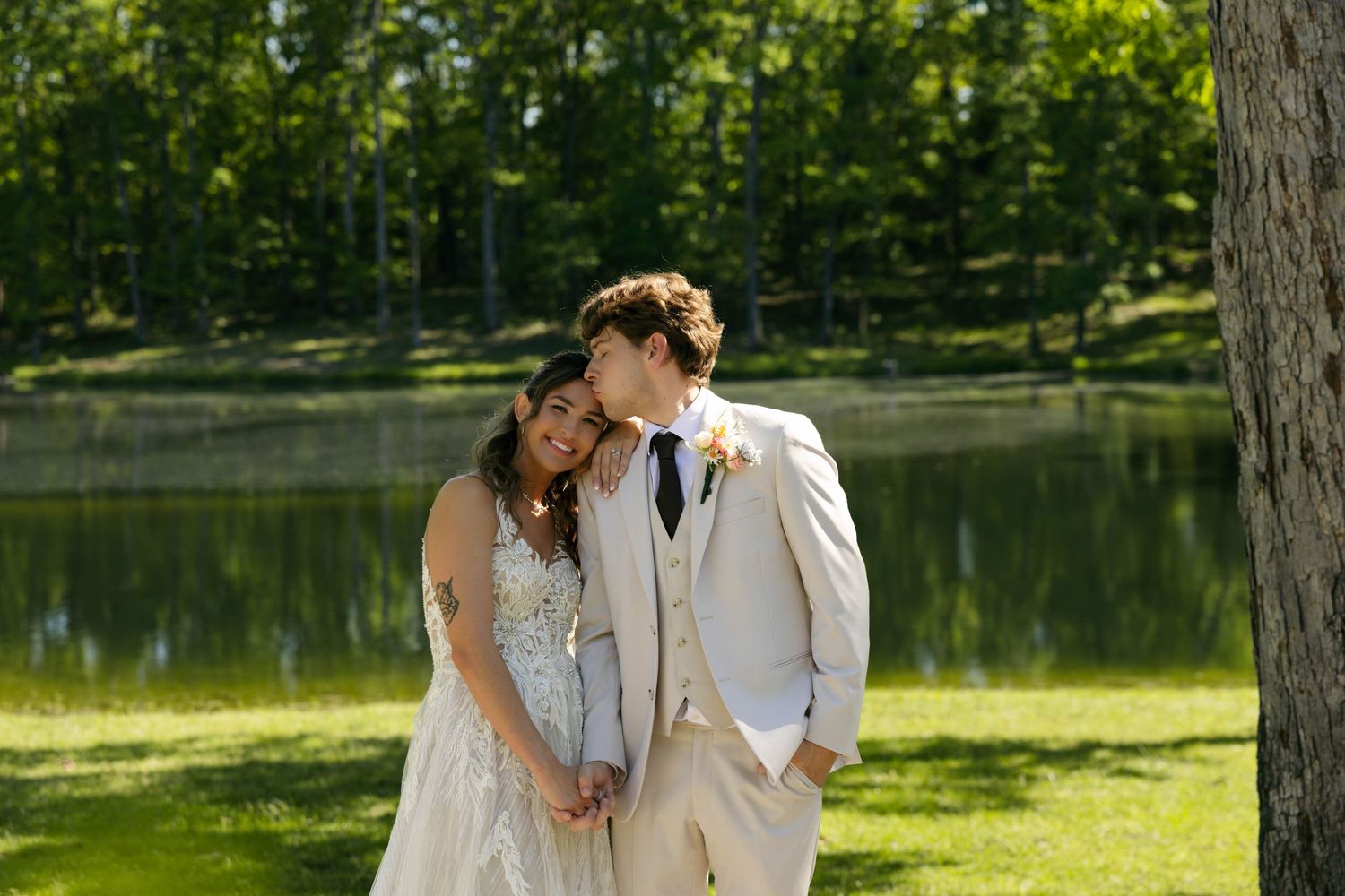 Bride and groom share a tender moment by a serene lake surrounded by lush green trees on their wedding day.