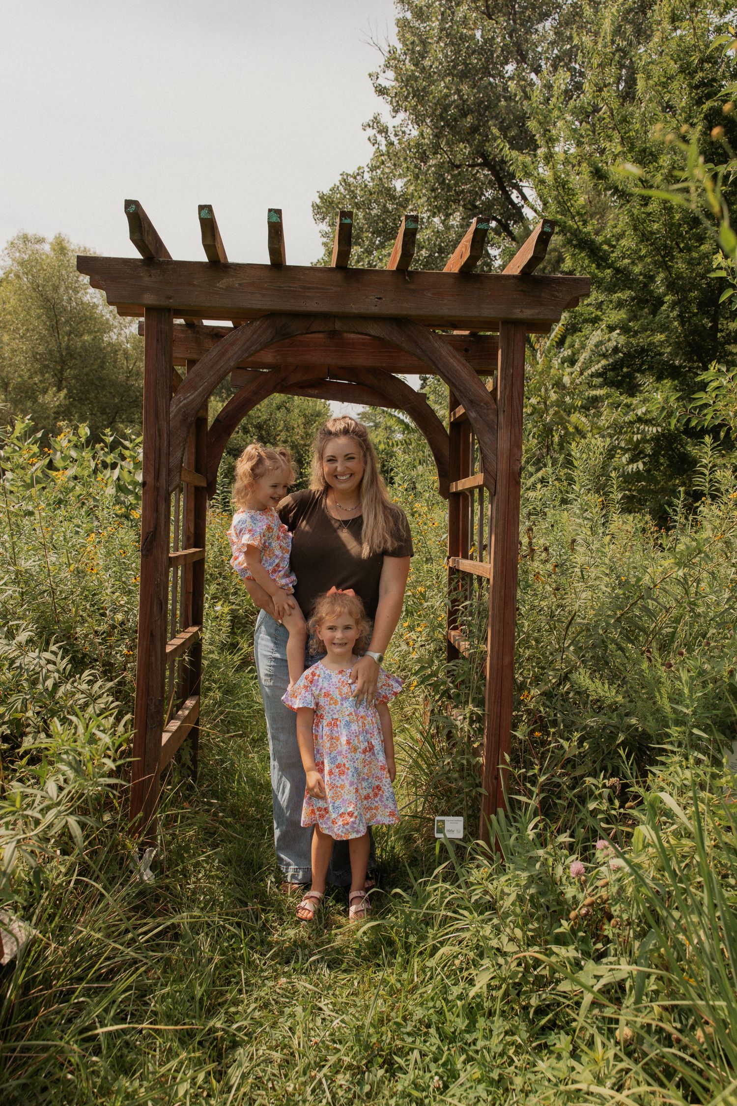 A rustic wooden arbor frames a family portrait in a natural garden setting.