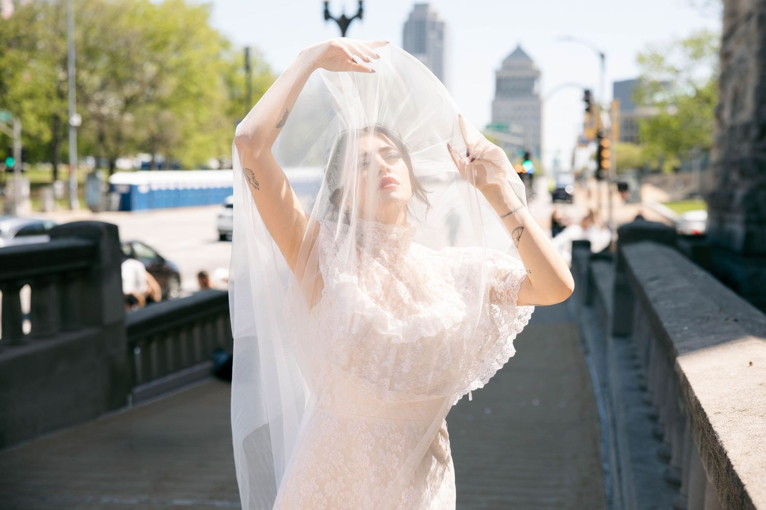 Bride in lace gown holds veil above head on sunny city bridge with skyline backdrop.