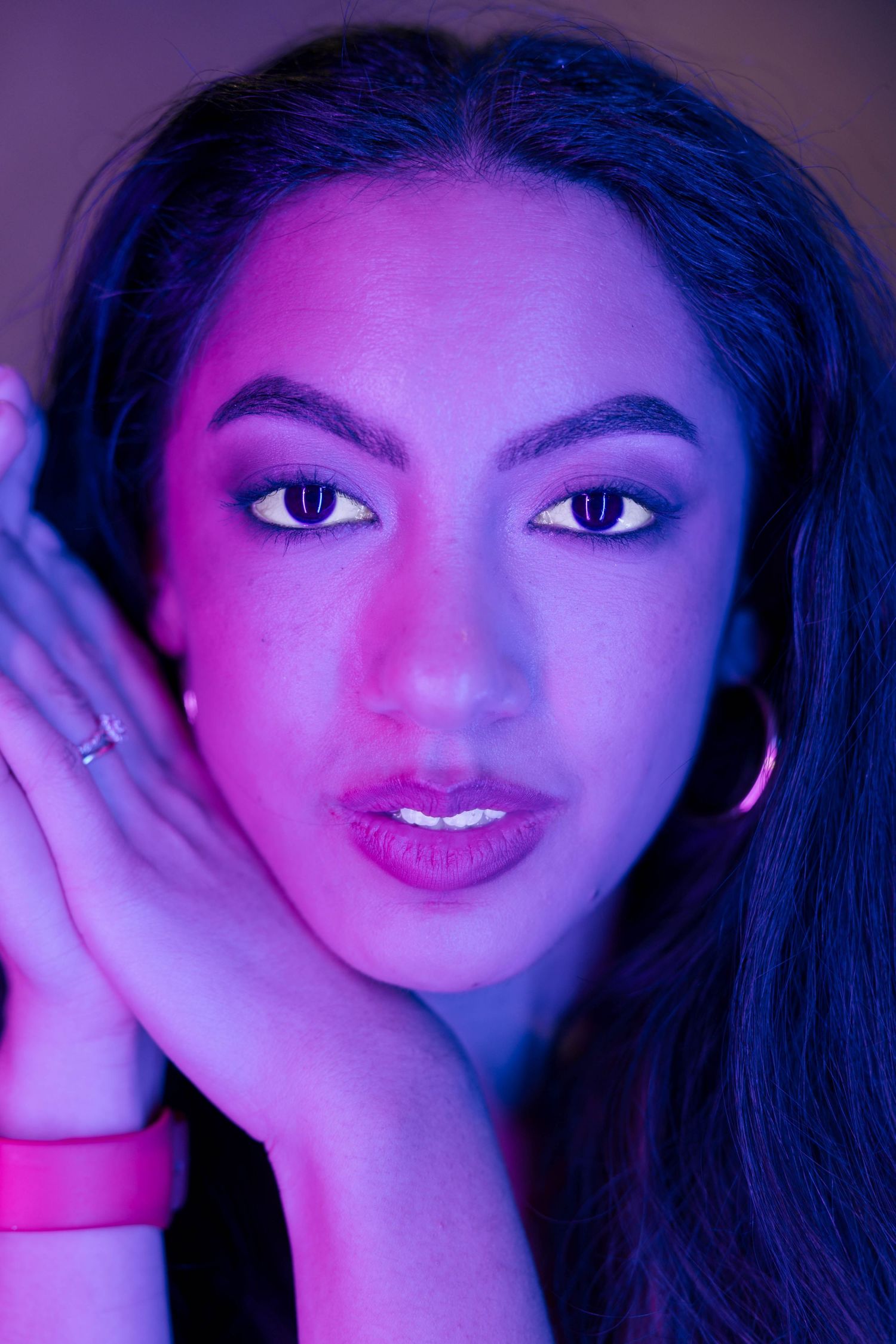 Close-up portrait of a young woman with bold eye makeup under purple and pink neon lighting.