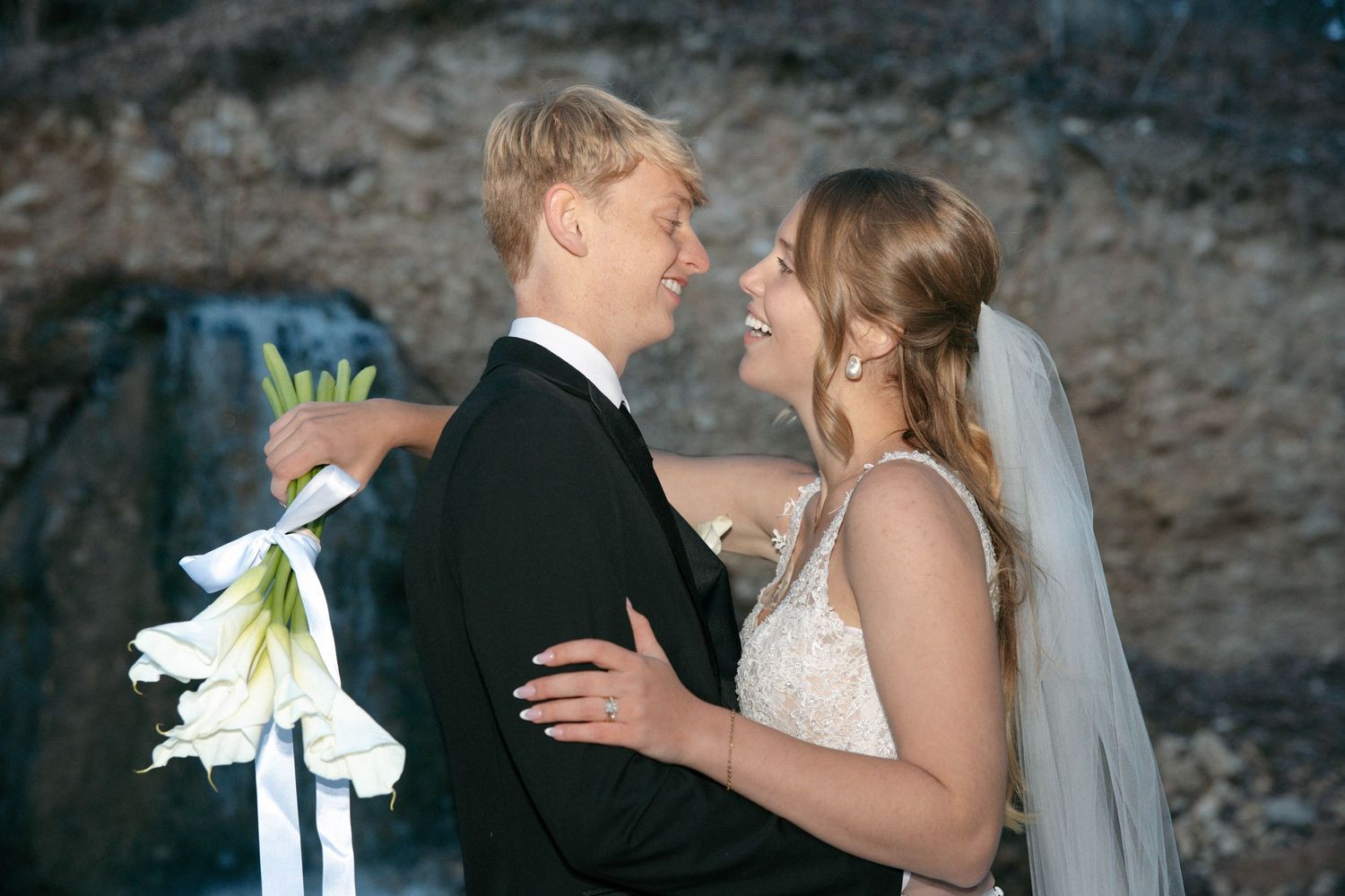 A bride and groom share a romantic moment outdoors near a waterfall, with the groom holding white lily bouquet.