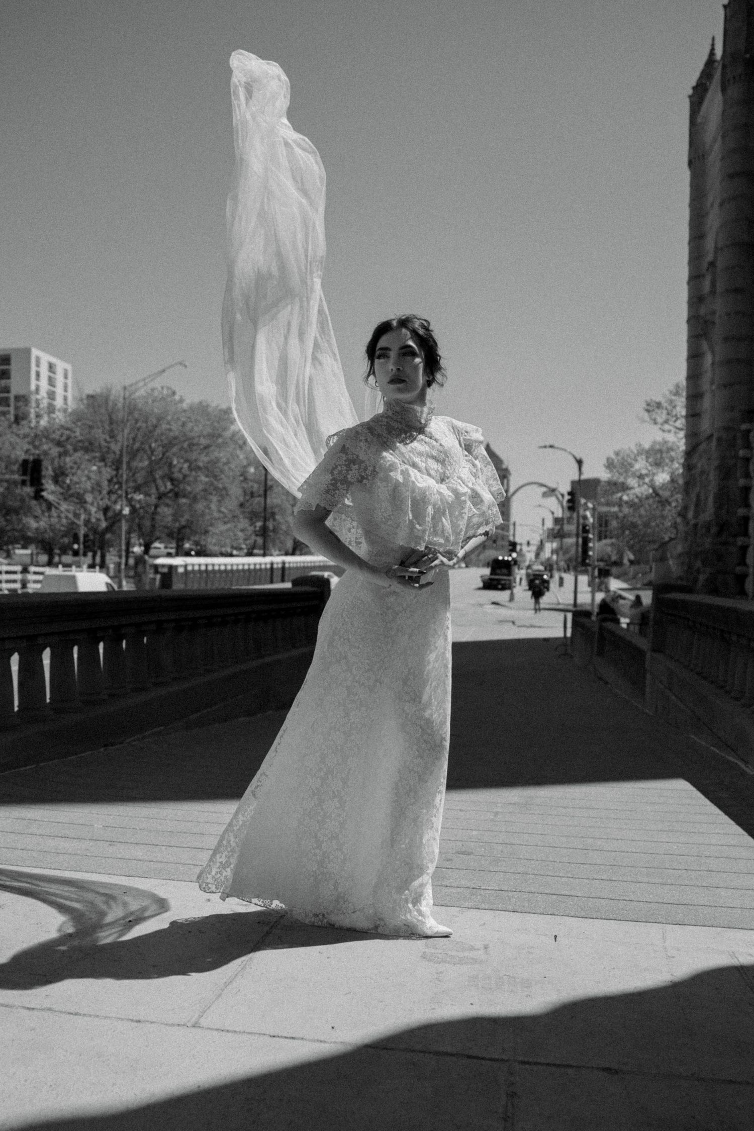 Elegant bride in lace dress with veil billowing upward on sunny city bridge, dramatic black and white.