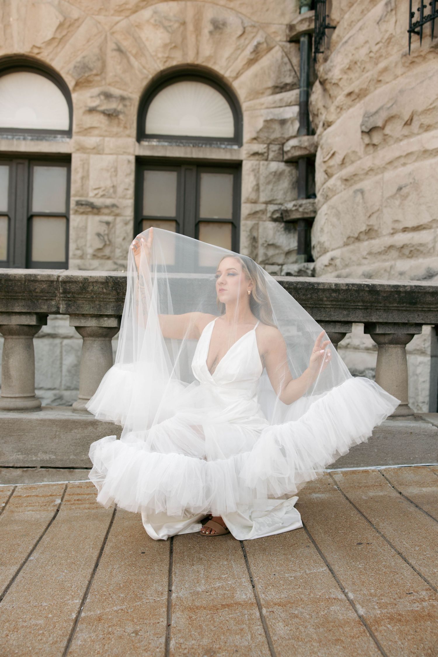 Bride in white tulle wedding dress and veil crouching on stone steps outside a historic building.
