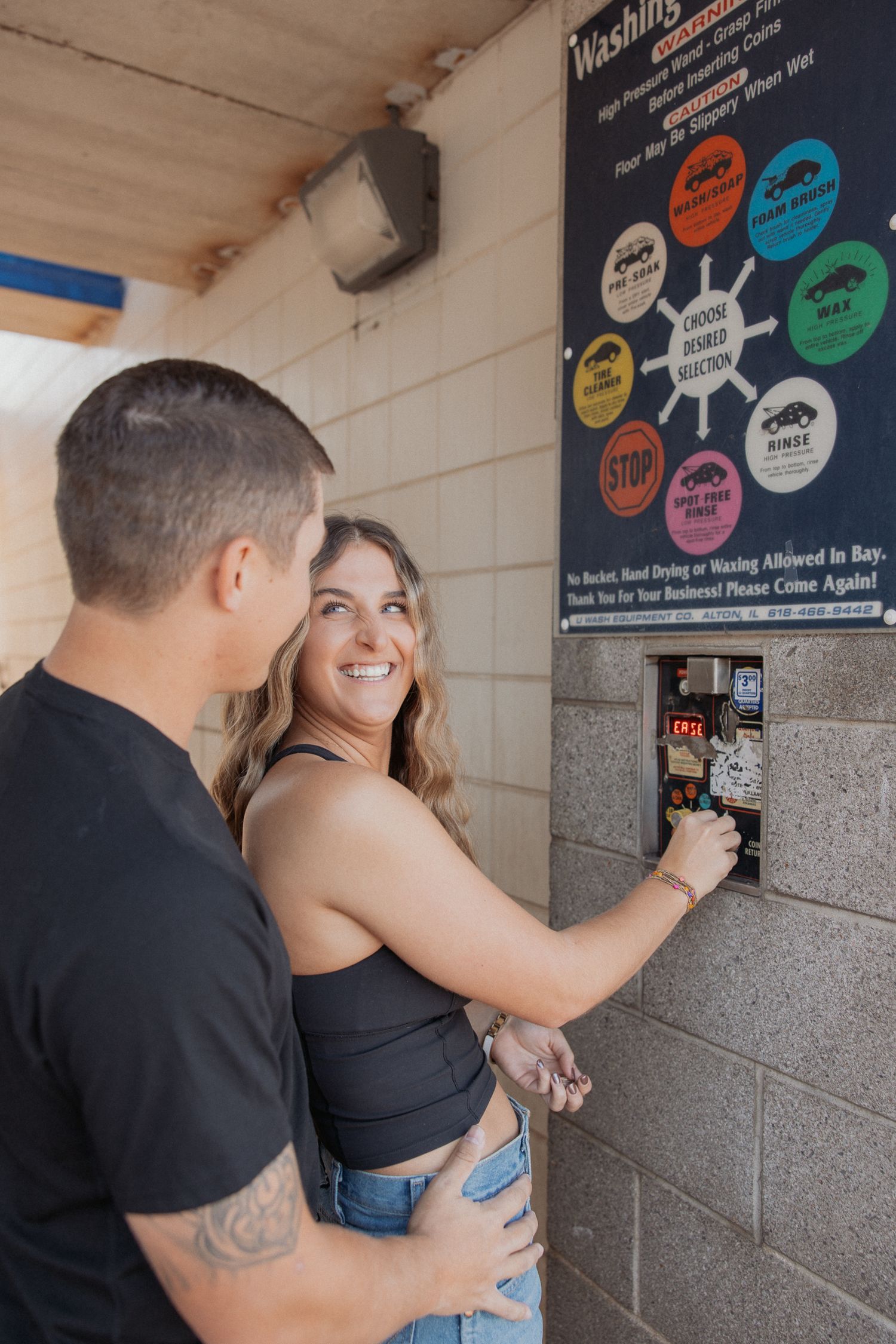 A couple playfully interacting near a wall with colorful signs and stickers.
