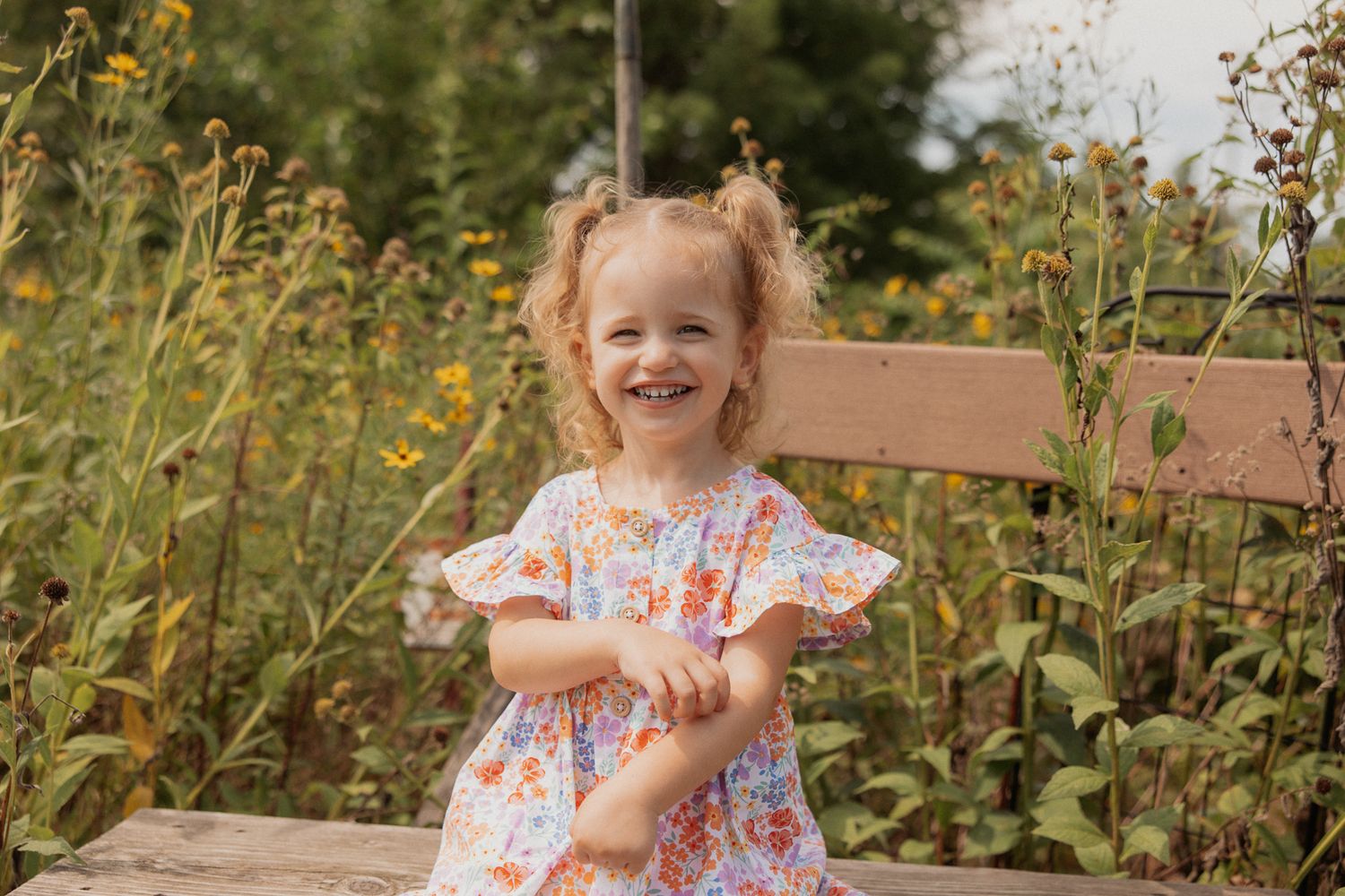 Child in floral dress laughing while sitting on wooden bench among wildflowers.