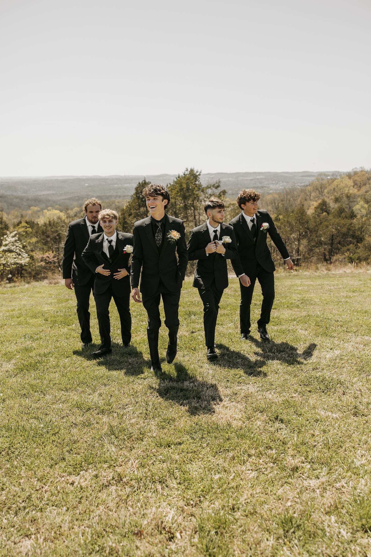 Groomsmen in black suits walk together across a hilltop meadow.