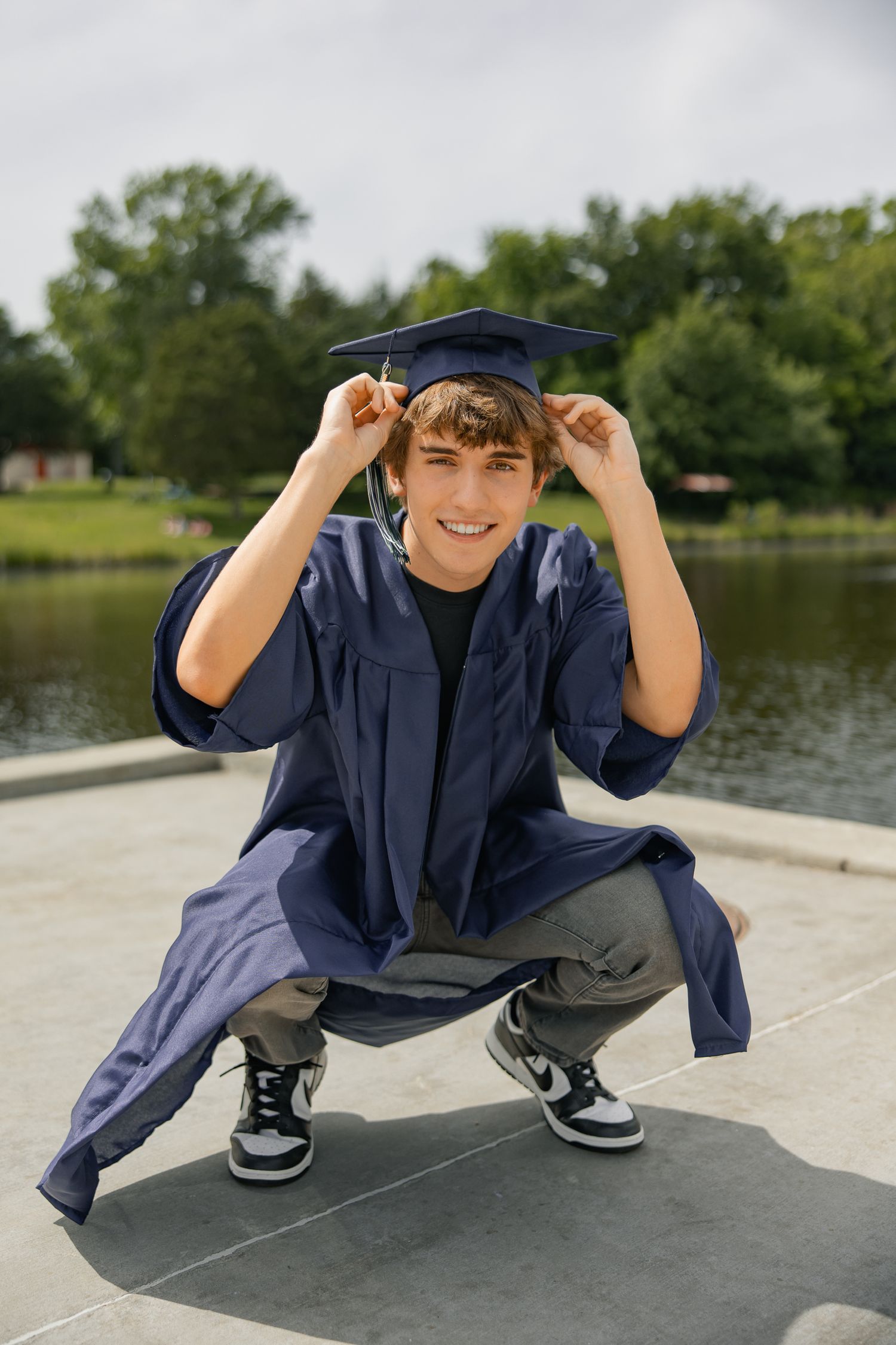 A graduate in blue regalia playfully adjusts their cap while crouching by a lake.
