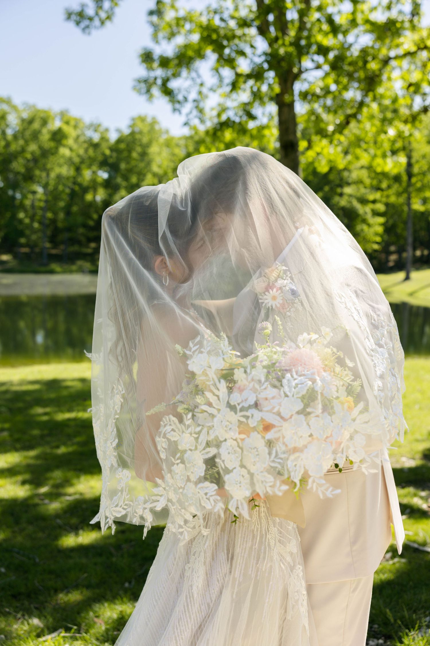 Bride and groom sharing a kiss under a lace wedding veil, holding a white floral bouquet in a lush green park.