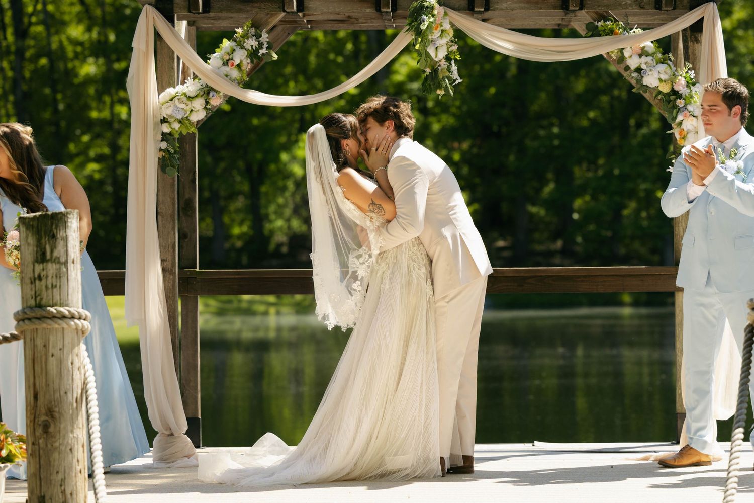 Bride and groom share first kiss on lakeside dock under floral arch at outdoor wedding ceremony.