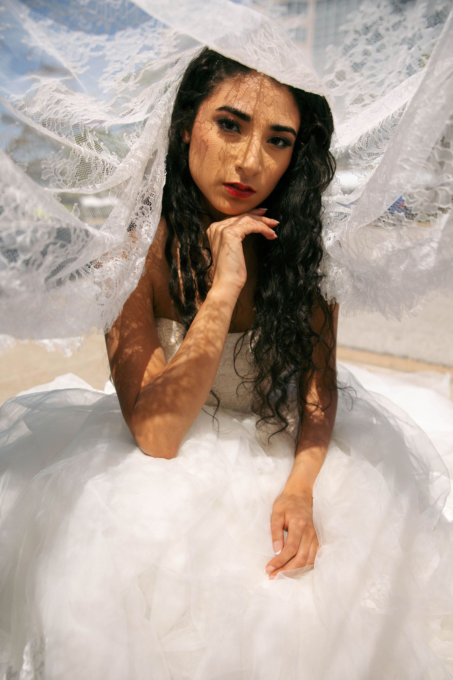 Bride in white ball gown and lace veil with dappled light on her face, posing elegantly outdoors.