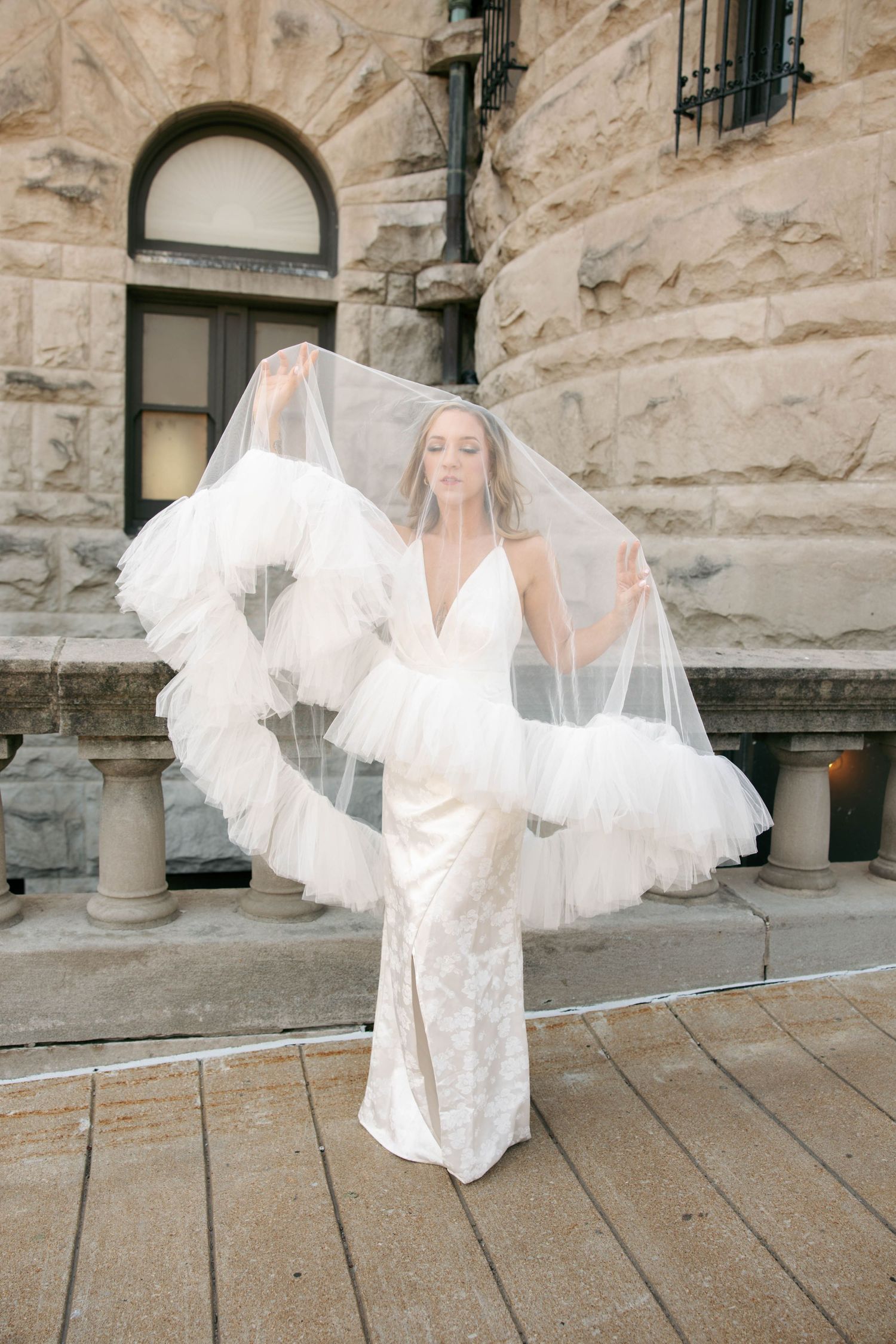 Bride in fitted lace gown holds flowing ruffled tulle veil outdoors against stone building.