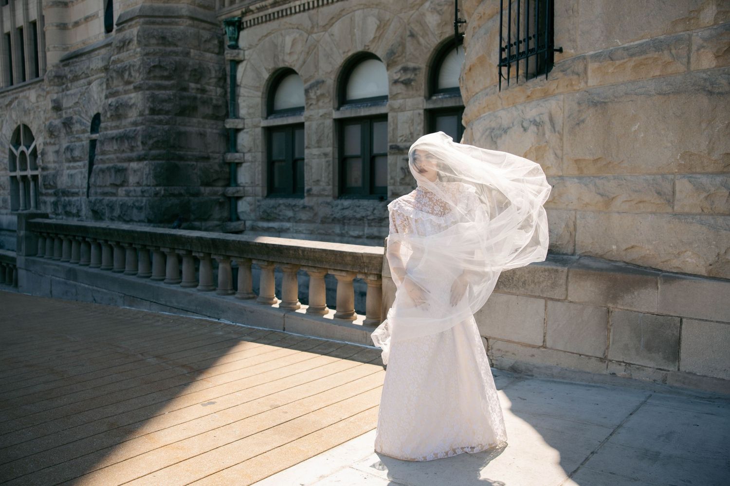 Bride in white wedding dress and flowing veil caught by wind, posing outdoors near historic stone building.
