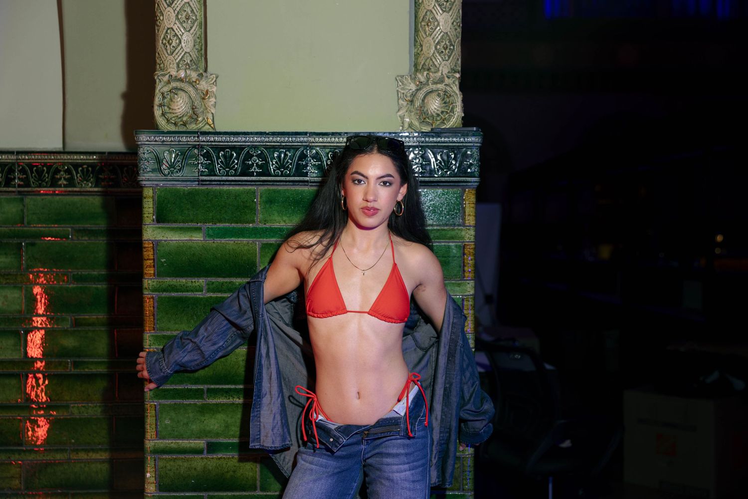 Woman in red bikini top, open jacket, and jeans posing in a dimly lit room.