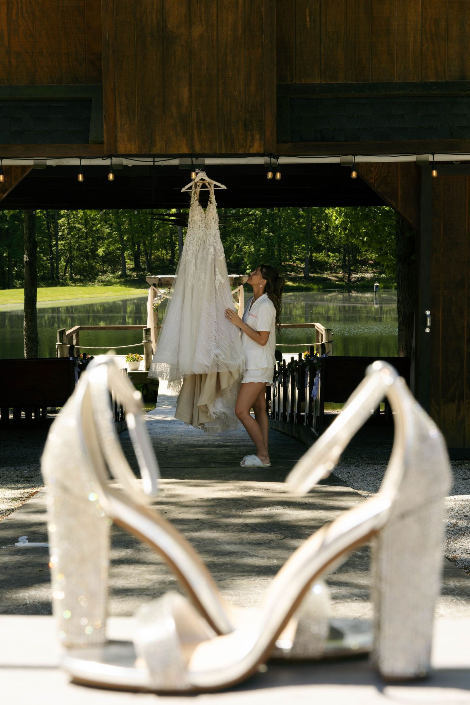 Bride examining her wedding dress hung outdoors, with sparkly white heels in the foreground at a rustic venue.