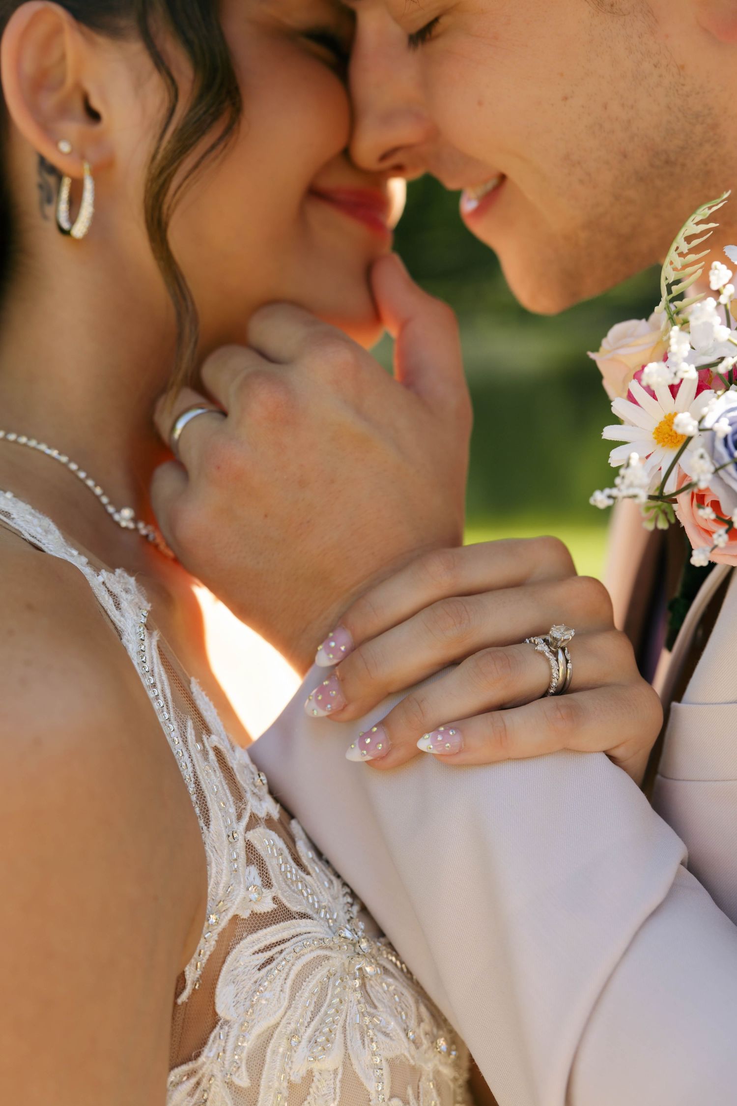 Close-up of bride and groom almost kissing, showing wedding rings, lace dress, boutonniere, and pearl necklace in golden sunlight.