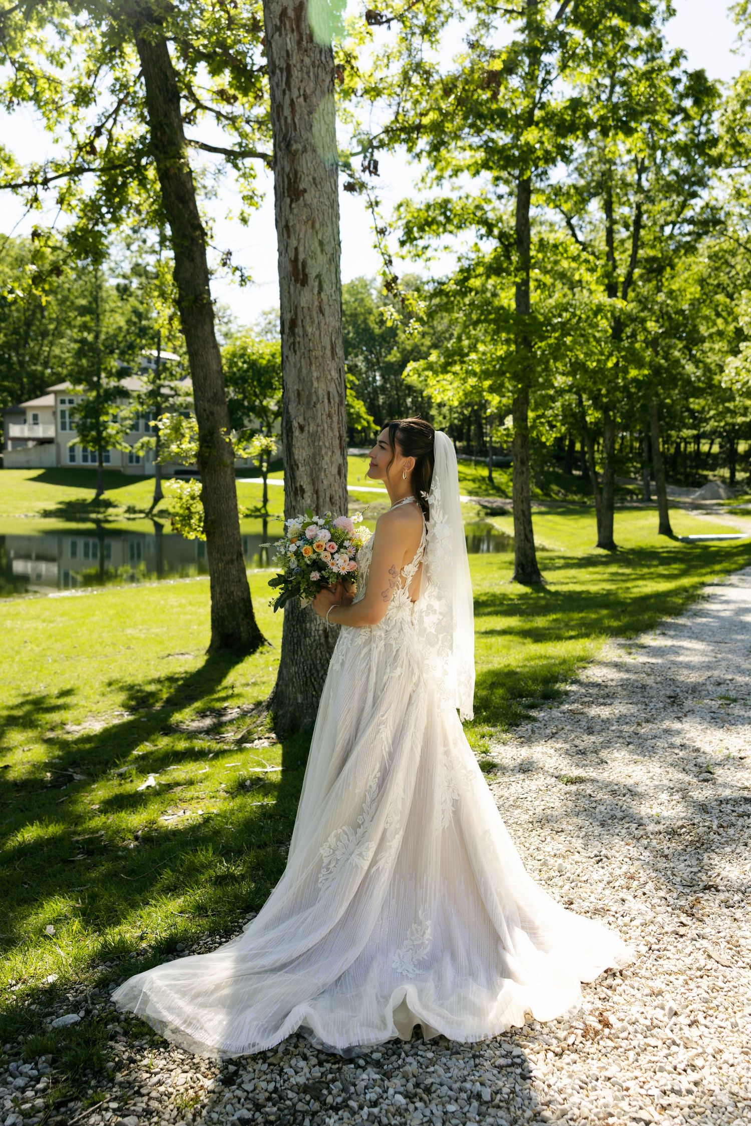 Bride in white lace gown with veil holds bouquet beside tree in sunlit green park.