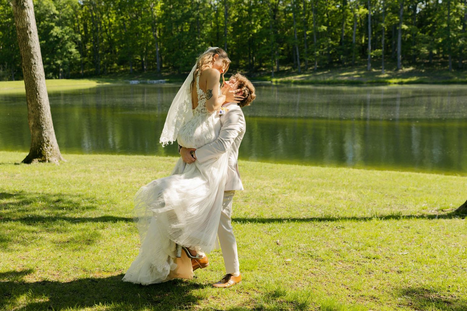 Bride and groom sharing a romantic kiss on green lawn beside a serene pond on their wedding day.