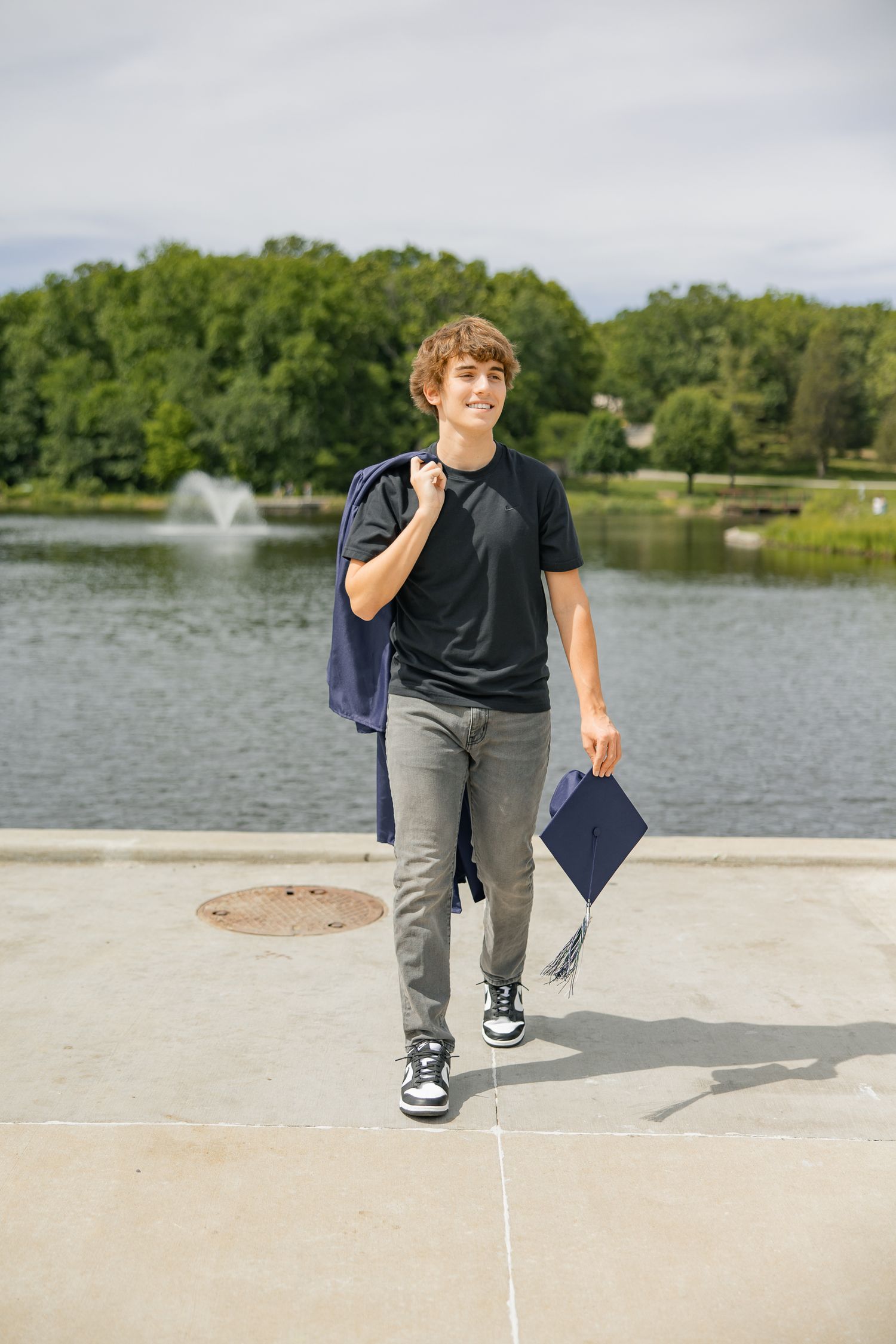 A graduate walks along a lakeside path carrying a diploma and wearing casual attire.