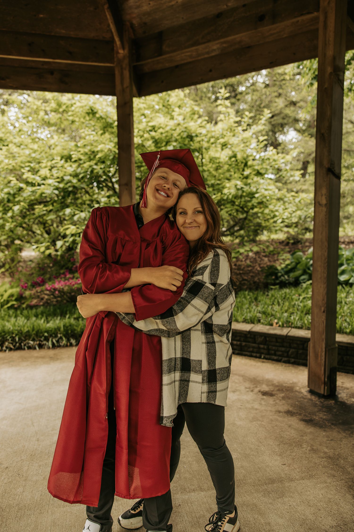A graduate in red academic regalia poses for celebratory photos under a covered walkway.