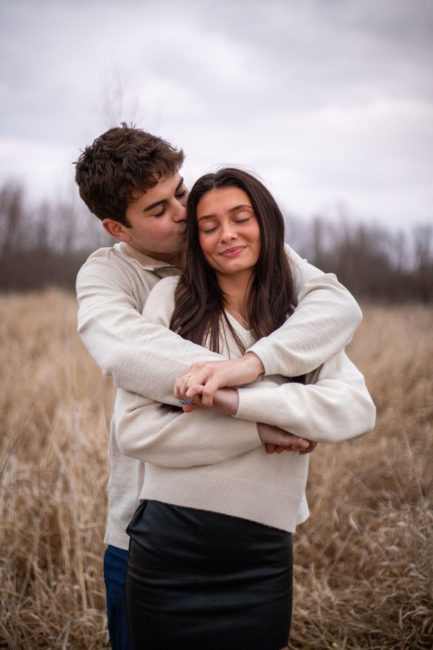 Romantic couple embraces in a field of tall dried grass on an overcast day.