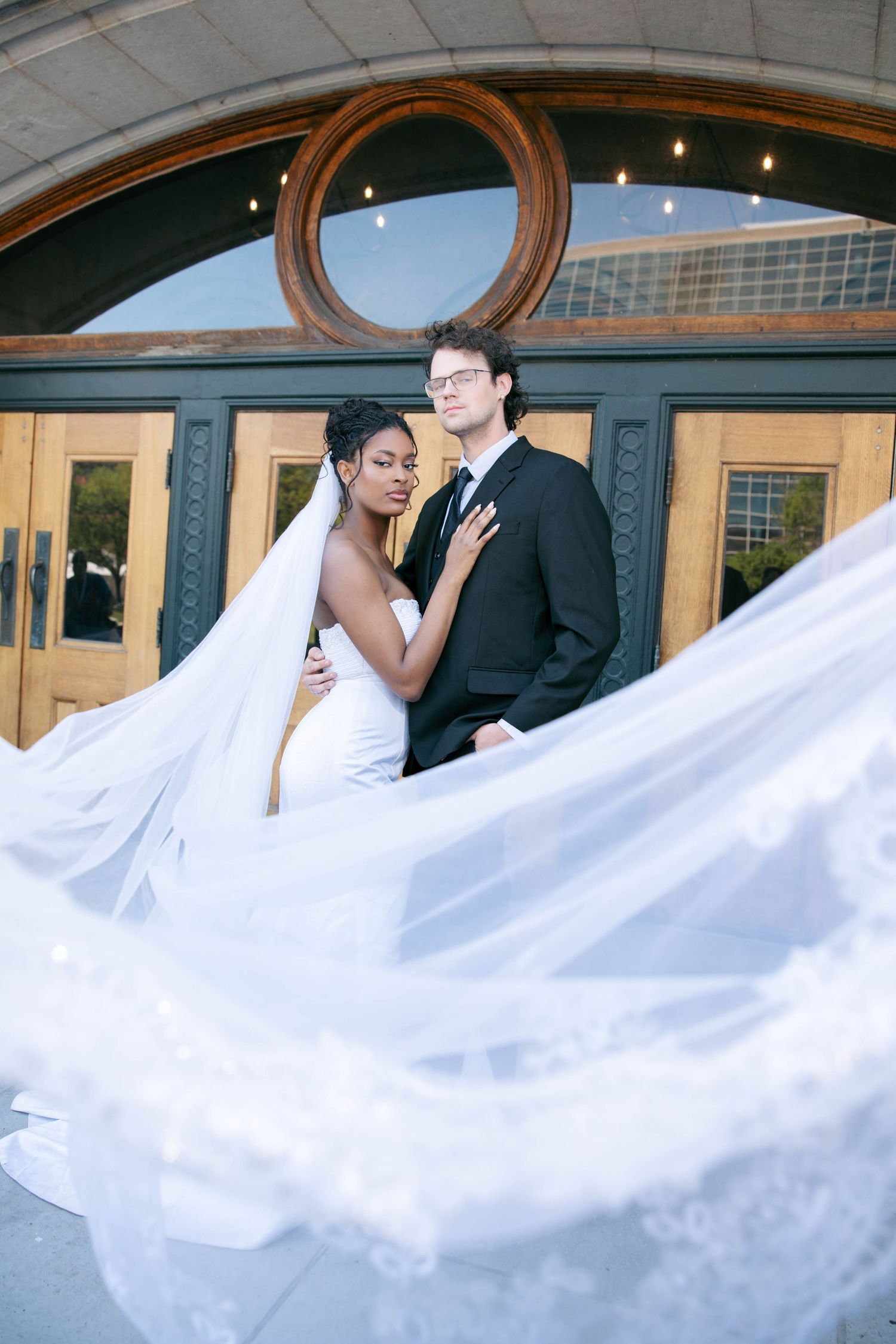 Interracial wedding couple embracing outside venue doors with bride's veil dramatically flowing in wind.
