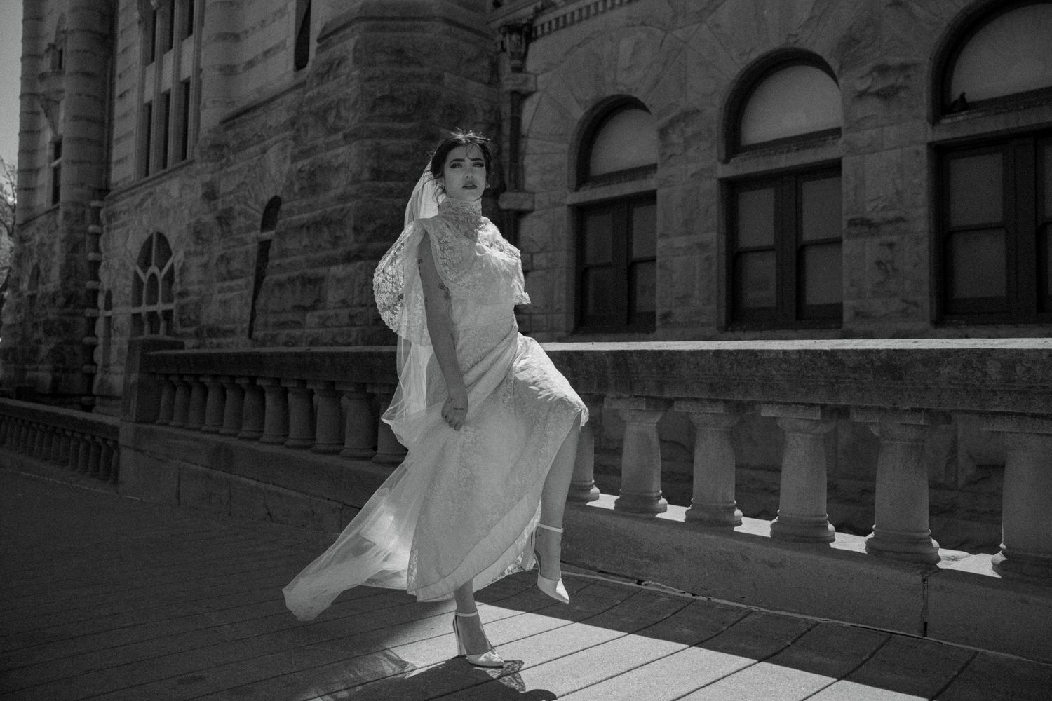 Black and white photo of a bride in a flowing white gown and veil walking on a stone balcony.