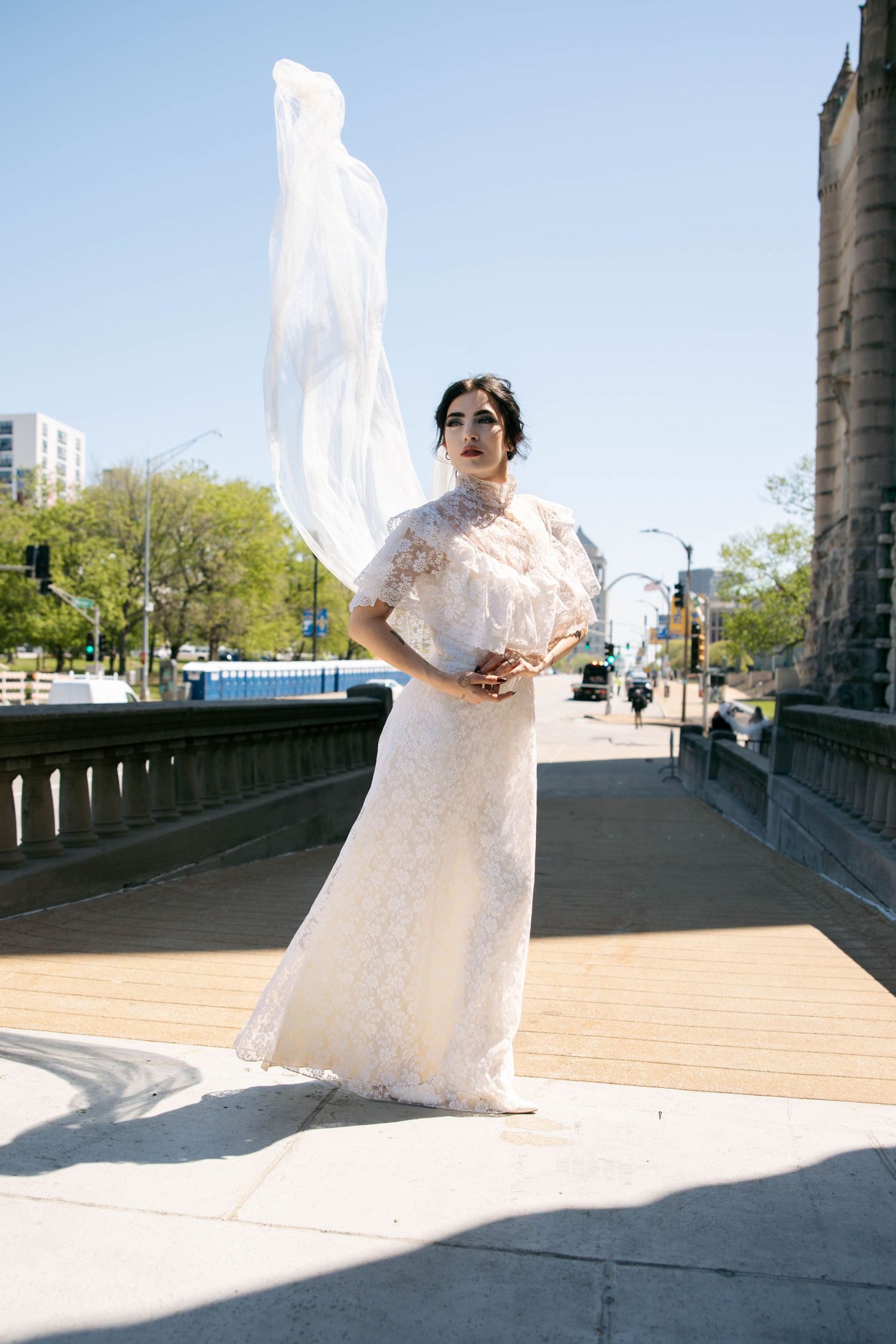 Bride in white lace gown standing on urban bridge with veil dramatically flowing in the wind.