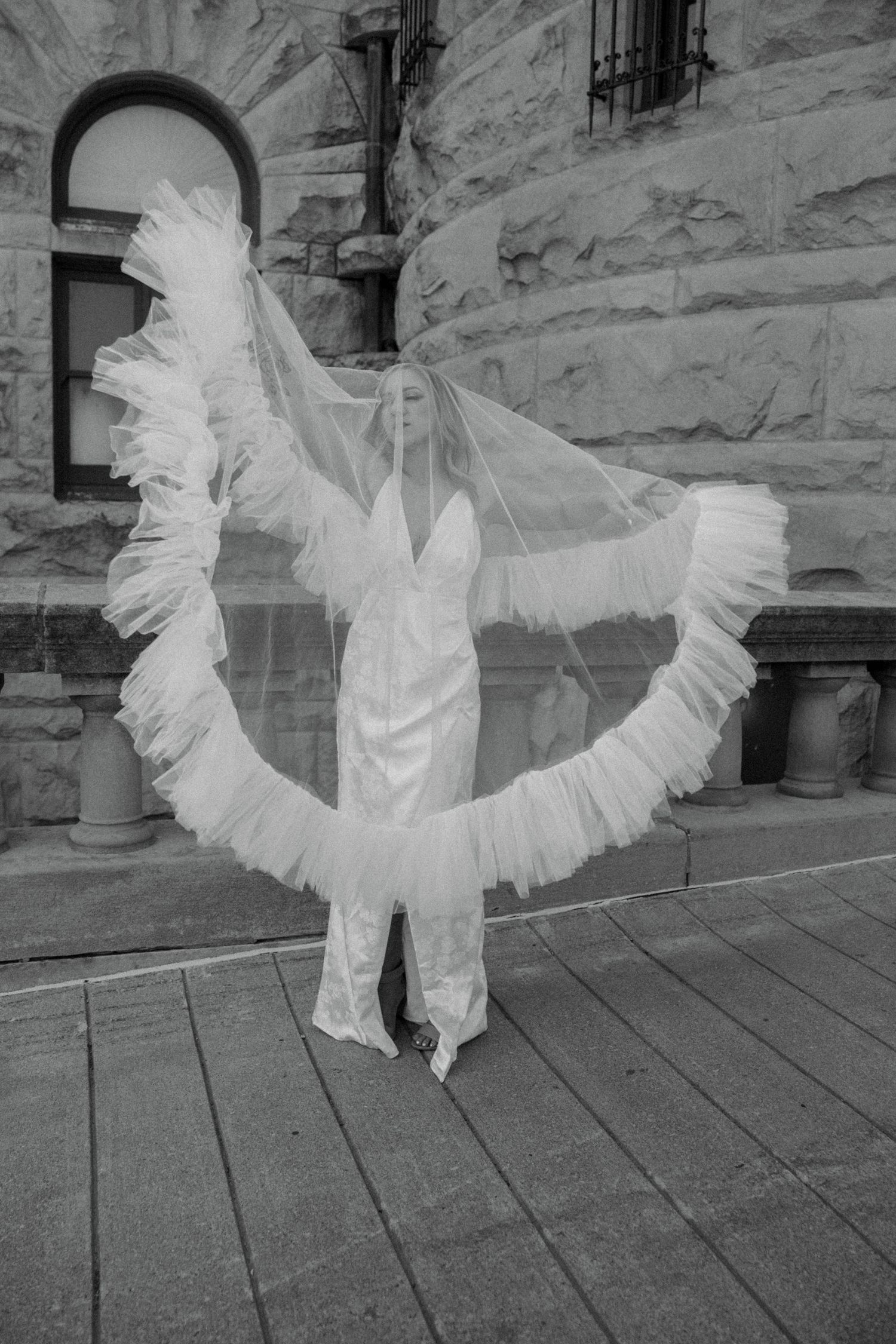 Black and white photo of bride in lace gown with dramatic ruffled veil spread wide outdoors.