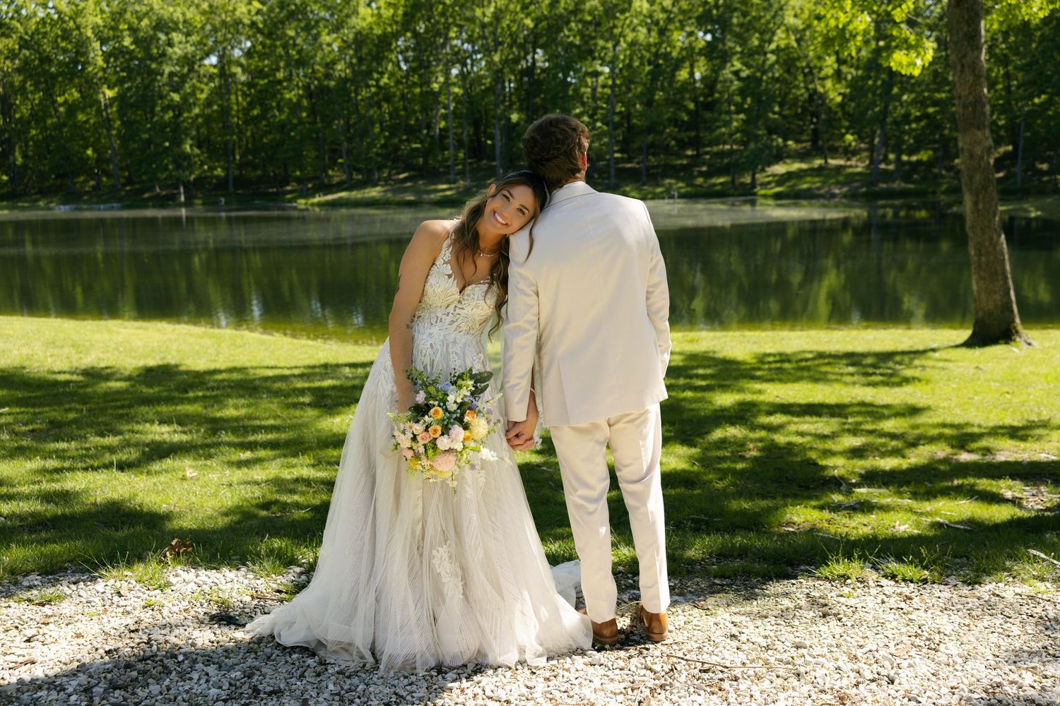 Bride in white gown holding bouquet and groom in tan suit hold hands near a serene pond surrounded by lush green trees.