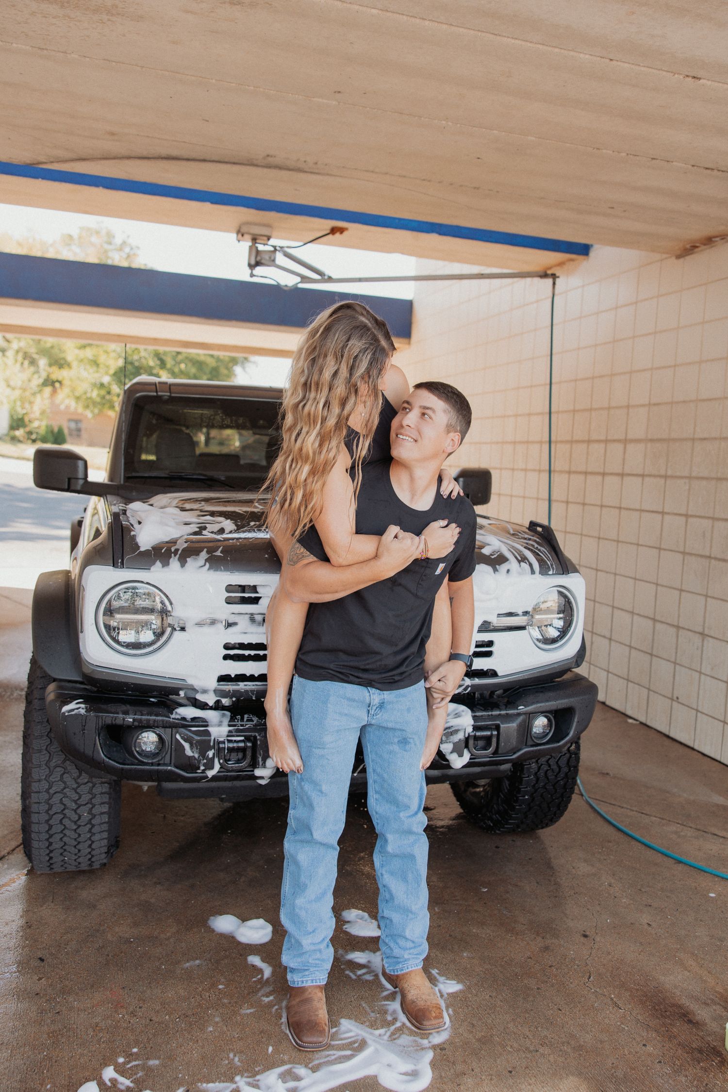 Two people share a tender moment while standing in front of a silver truck in a covered parking space.
