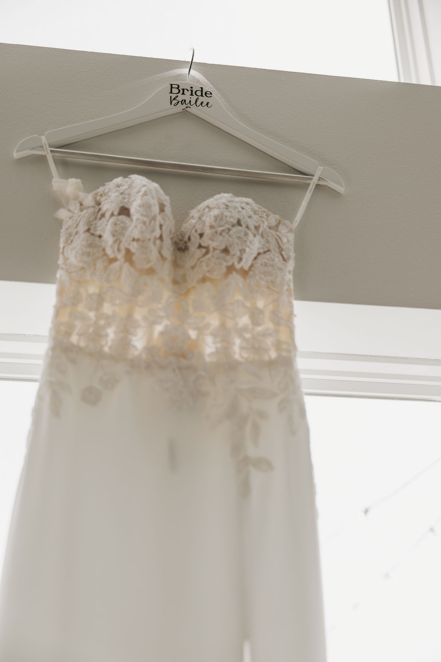 White lace wedding dress with sweetheart neckline hanging on bride-to-be hanger against light wall.