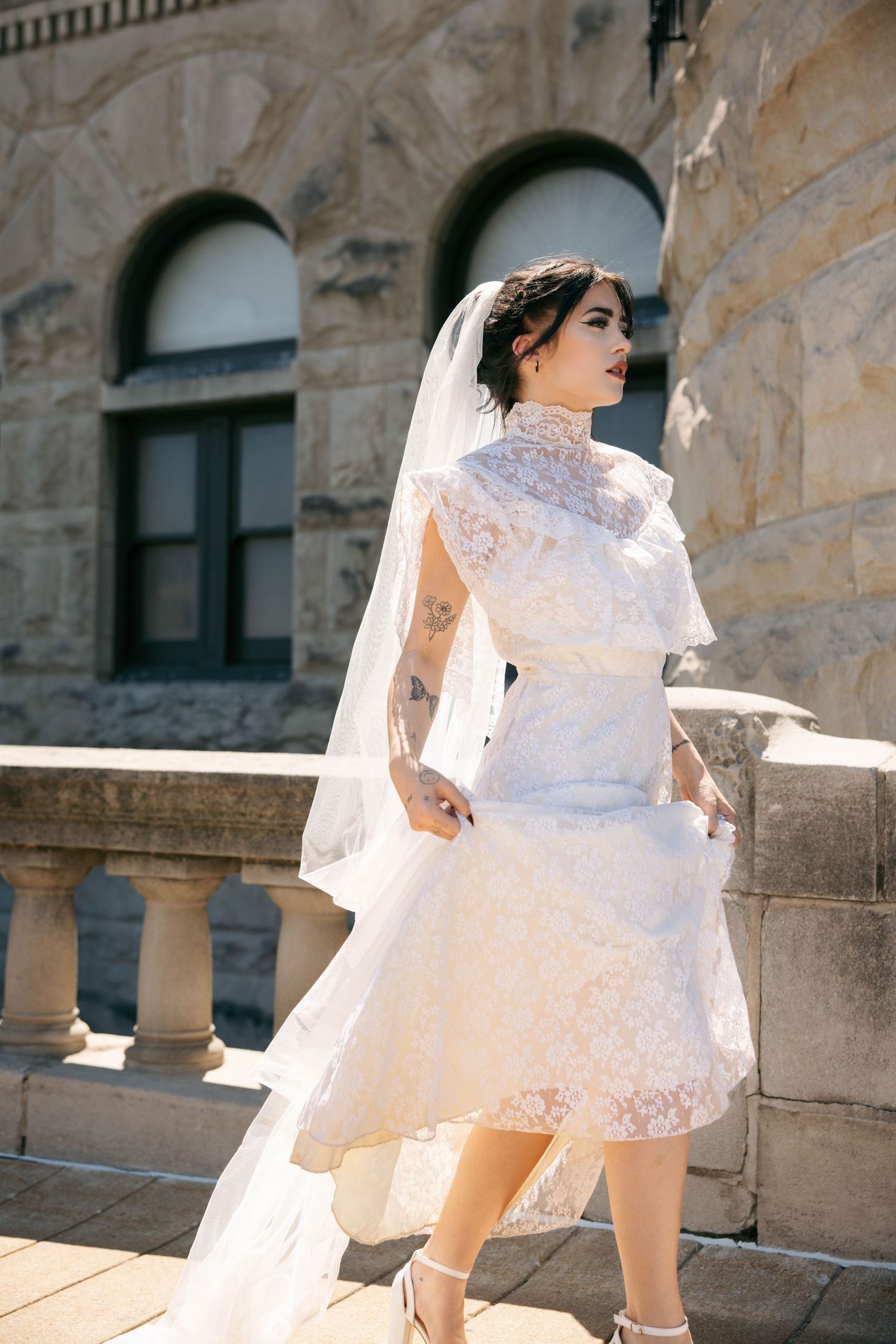 Elegant bride in knee-length lace wedding dress and veil posing on stone balcony in sunlight.