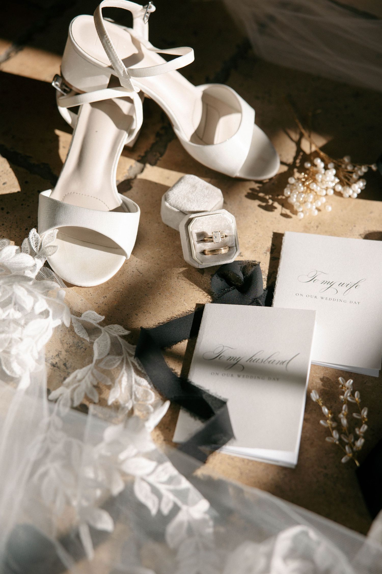 White bridal heels, wedding rings, vow books, and floral accessories arranged elegantly on a textured surface.