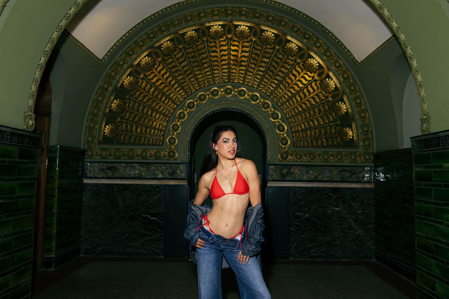 Woman in red bikini top and jeans posing in ornate arched alcove with gold mosaic tilework.