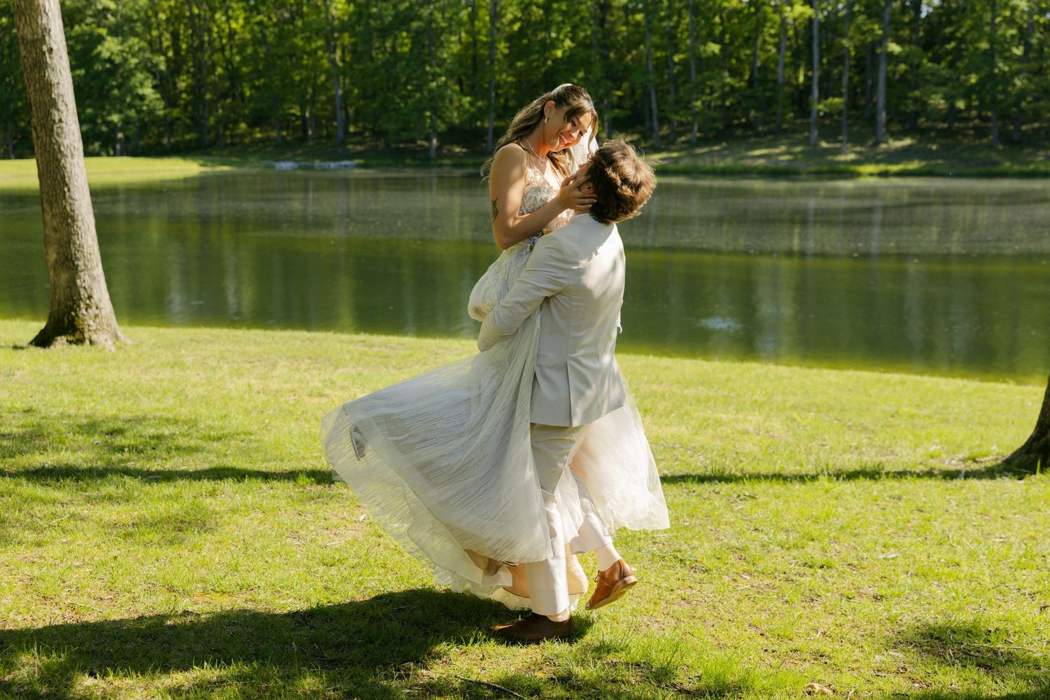 Couple in wedding attire dancing joyfully on green grass near a sunlit pond surrounded by trees.