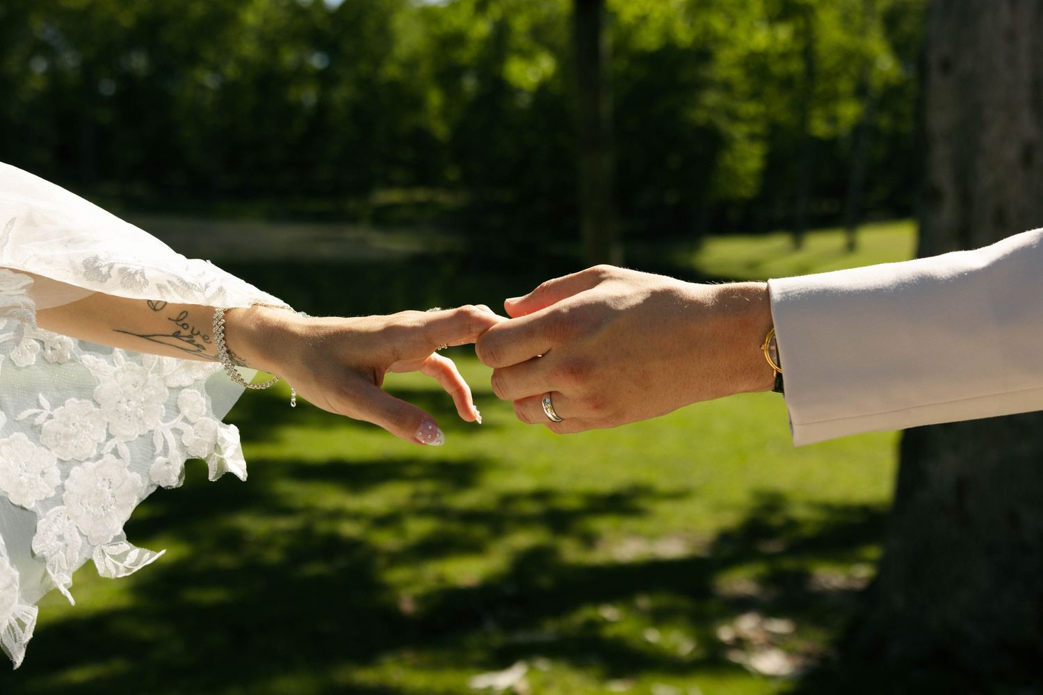 Bride and groom holding hands outdoors, showcasing wedding rings, with lush green trees in the background on a sunny day.